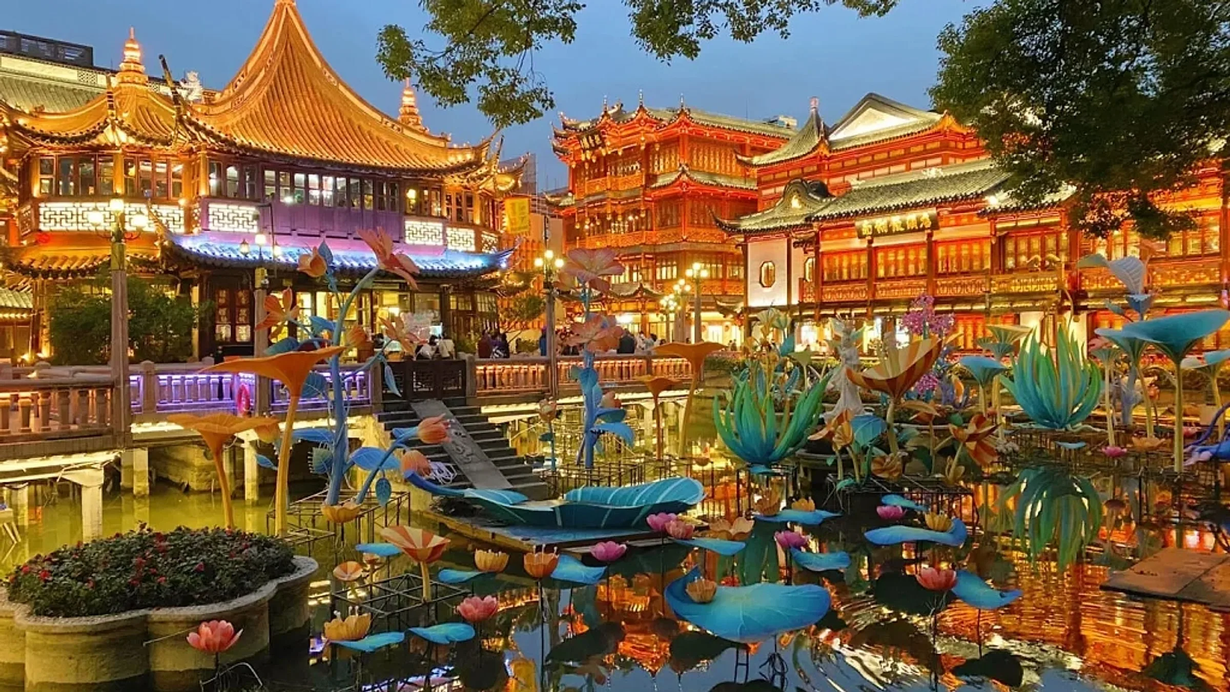 Yu Garden at night with traditional Chinese pavilions lit in orange and colorful lotus lanterns in the foreground pond
