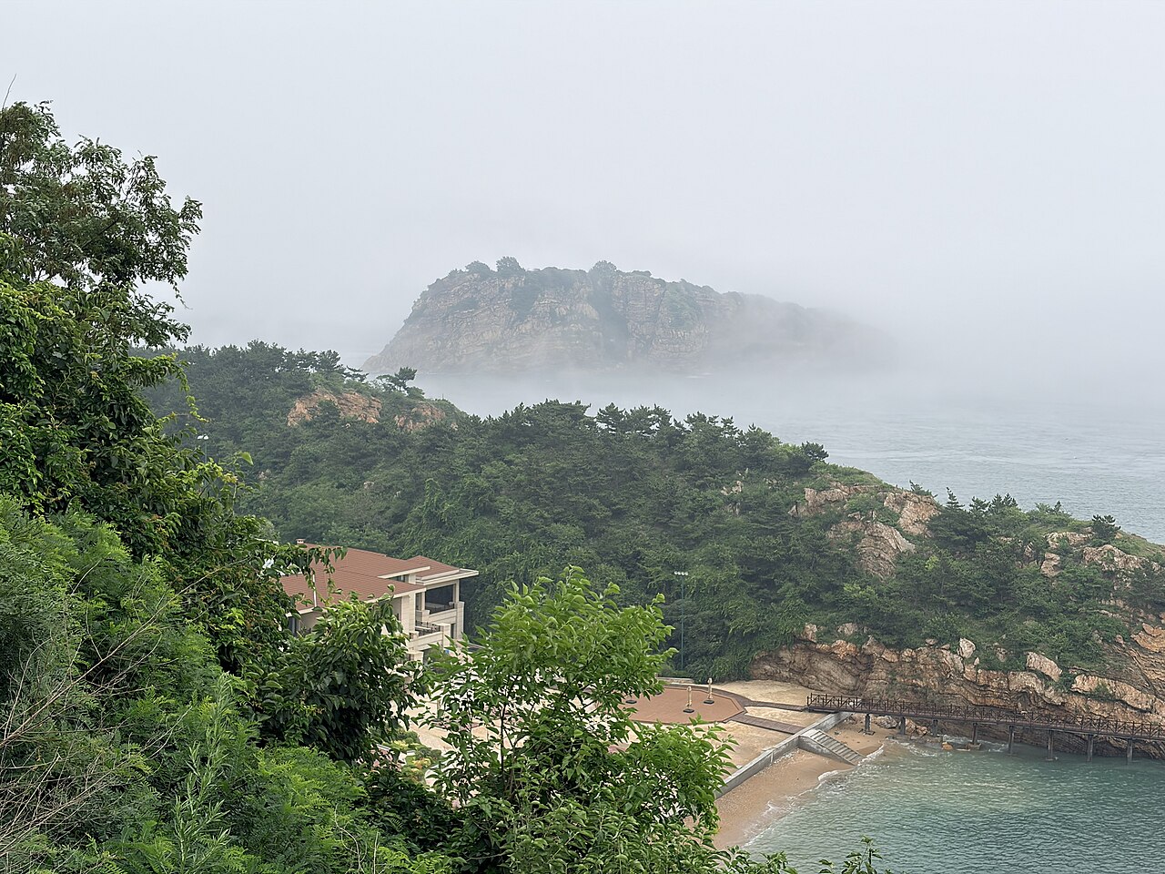 View of Bangchui Island, a small forested islet 600 m off the Dalian coast, with the white-sand beach in the foreground, the wooden pestle-shaped rock on the islet's peak visible, and the Yellow Sea and the Dalian coastal hills in the background.