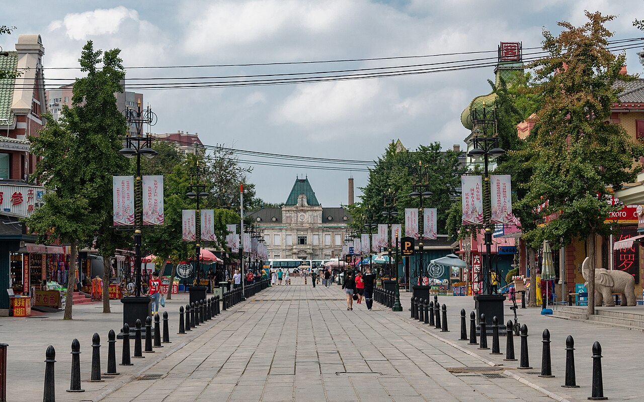 A row of restored 1900s Russian baroque buildings in warm yellow and cream tones along Russian Street in central Dalian, with a stone bridge crossing a small canal in the foreground, late afternoon golden light, and a few visitors walking the pedestrian street.