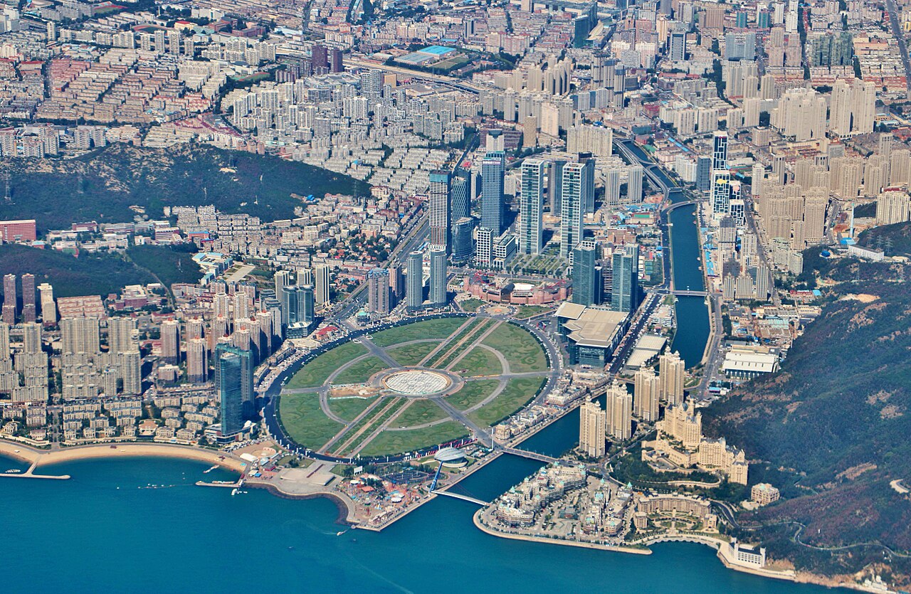 Aerial view of Xinghai Square and the surrounding skyline of Dalian at twilight, with the Century Sculpture's 19.9-m open book illuminated, the sea in the distance, and the Yifang Center skyscraper lit up in the background.