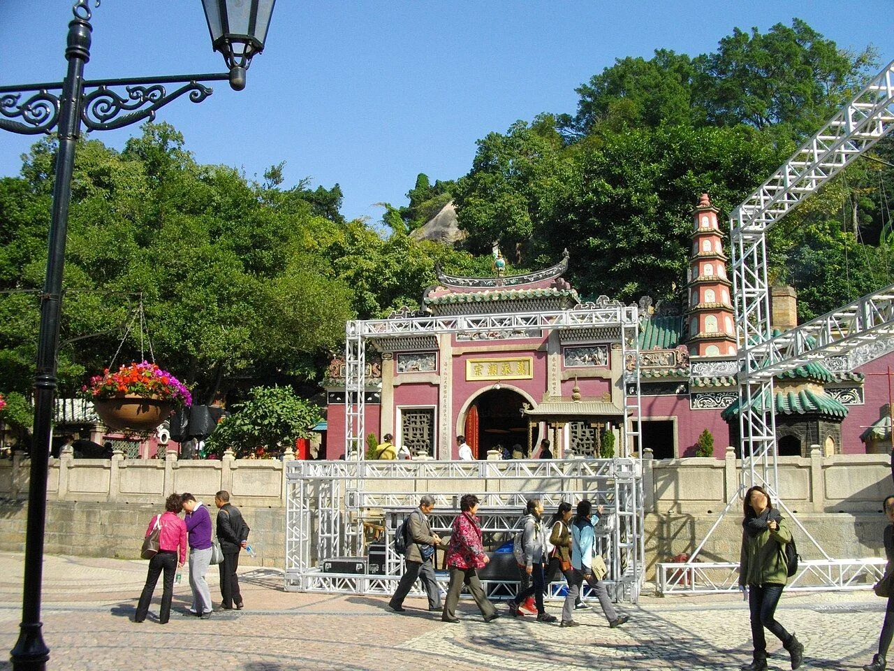 The A-Ma Temple entrance courtyard in Macau, with visitors, red lanterns, the carved stone guardian figures of the Hall of the Mountain, and the minaret-style pavilion of the Goddess in the background