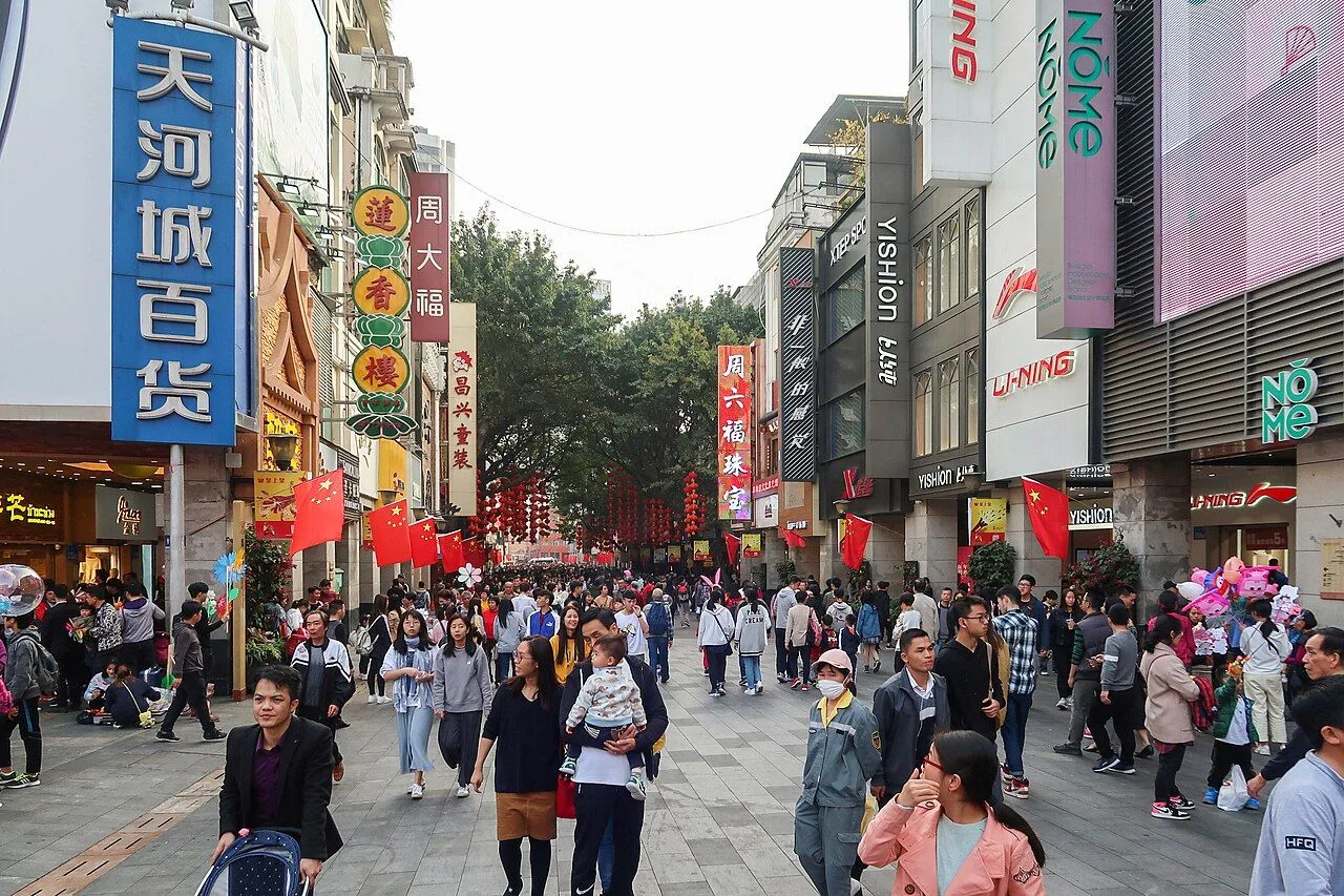 Crowded Beijing Road pedestrian street in Guangzhou on a typical day, shoppers walking between 1930s arcade buildings with Chinese flag decorations and red lanterns hanging overhead