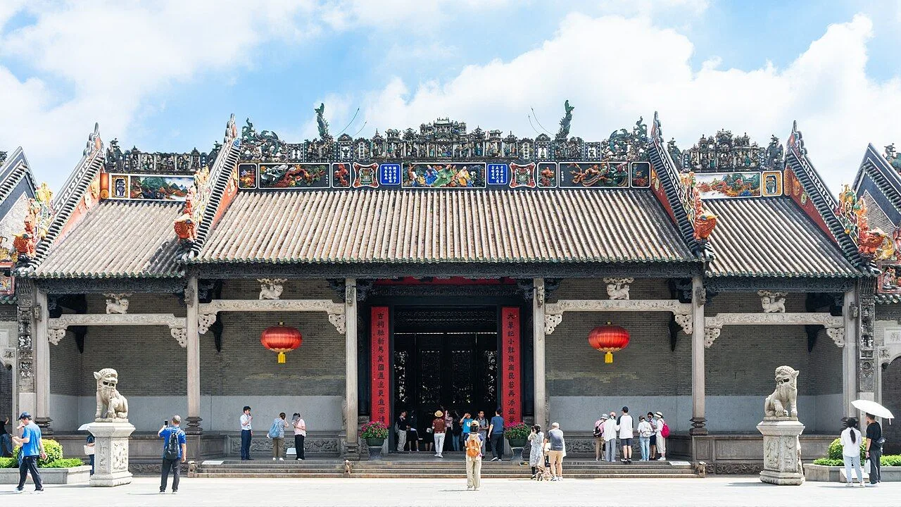 The main entrance of the Chen Clan Ancestral Hall in Guangzhou — the 1894 Qing-dynasty Lingnan-style facade with both stone guardian lions, the characters 陳氏書院 on the red signboard, ceramic roof-ridge sculptures, and visitors entering