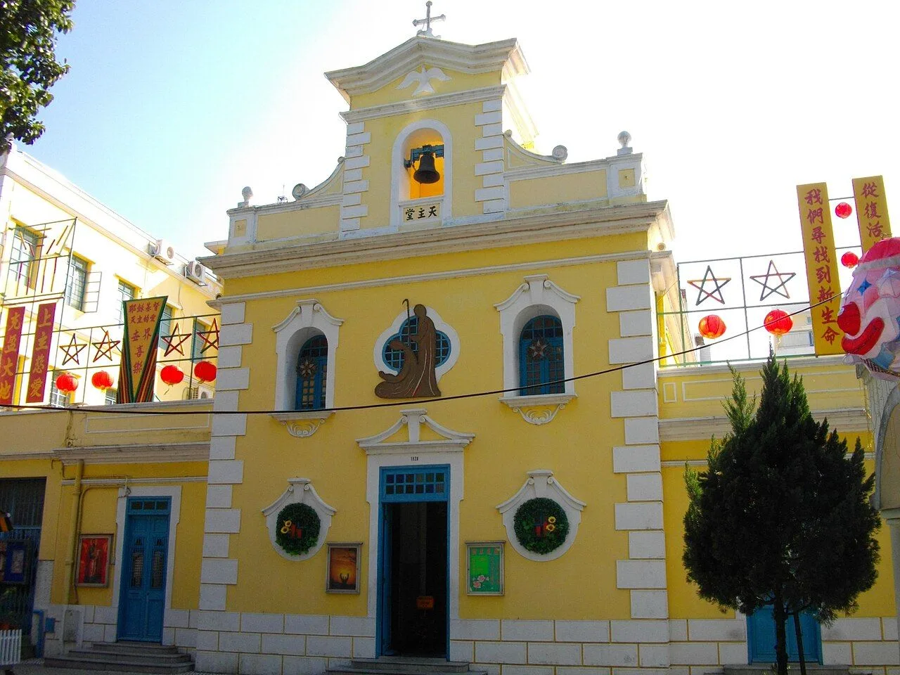 The yellow St. Francis Xavier Chapel in Coloane Village, Macau — the 17th-century Catholic chapel with white trim, blue doors, Christmas wreaths, and Chinese New Year banners reflecting the hybrid Chinese-Portuguese heritage of the village