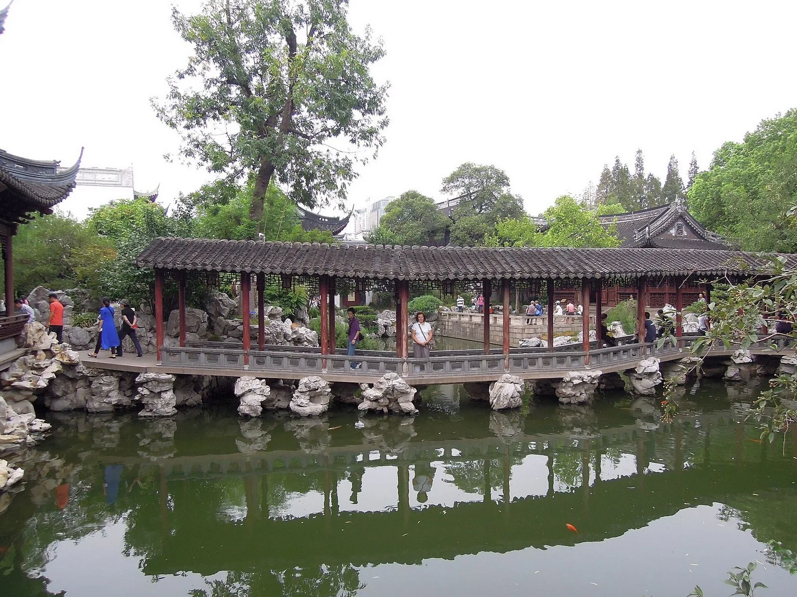 The famous zigzag covered bridge in Yu Garden, Shanghai — a long covered walkway with traditional gray-tile roof supported by red wooden columns, crossing the central pond on stone piers, with several visitors walking along it, a large tree rising behind, and the garden's traditional upturned-eave pavilions visible in the background. The zigzag design was traditionally believed to keep evil spirits from crossing water