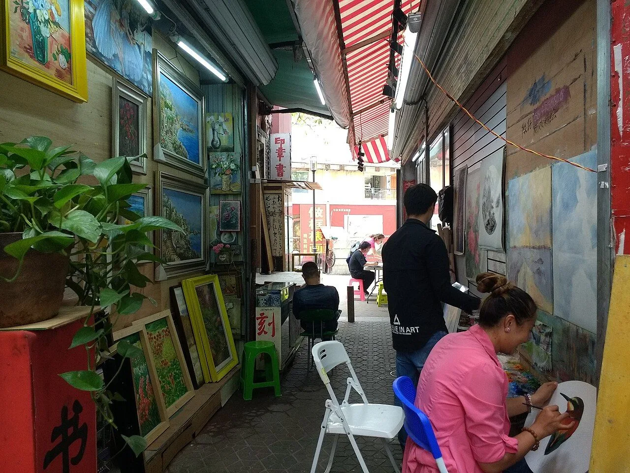 Inside a working studio in Dafen Oil Painting Village, Shenzhen, showing two artists (one in pink) seated and hand-painting oils on canvas, with framed finished paintings hung on the walls and along the narrow alleyway of the village