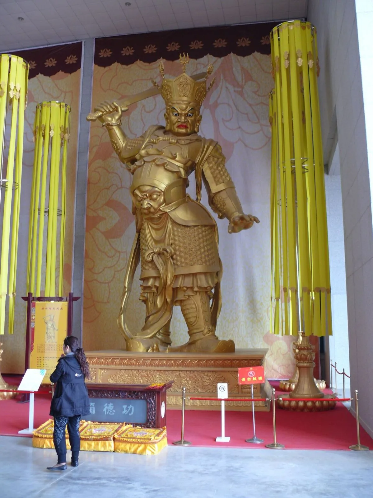 A massive golden Heavenly King (Virūḍhaka) guardian statue inside Famen Temple, holding a sword and standing in a protective pose, with a female visitor in a black coat reading the placard in front of the statue for scale
