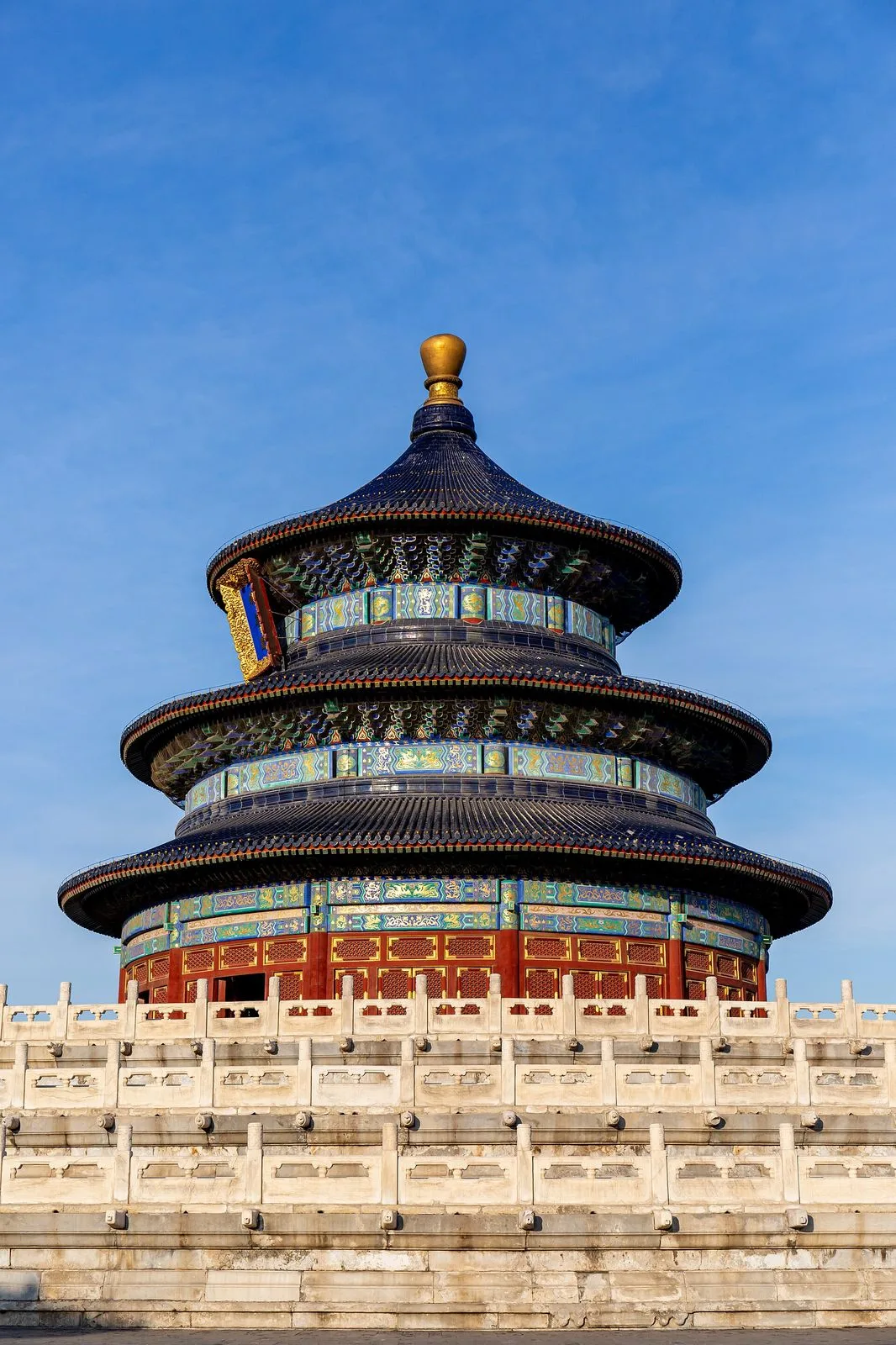 The Hall of Prayer for Good Harvests at the Temple of Heaven, viewed from below on a clear blue-sky day — the 38-meter tall triple-eaved circular building with deep blue glazed tiles, gold finial, and the white marble triple-tiered platform with carved balustrades at its base. The most iconic image of imperial Beijing, a UNESCO World Heritage Site since 1998