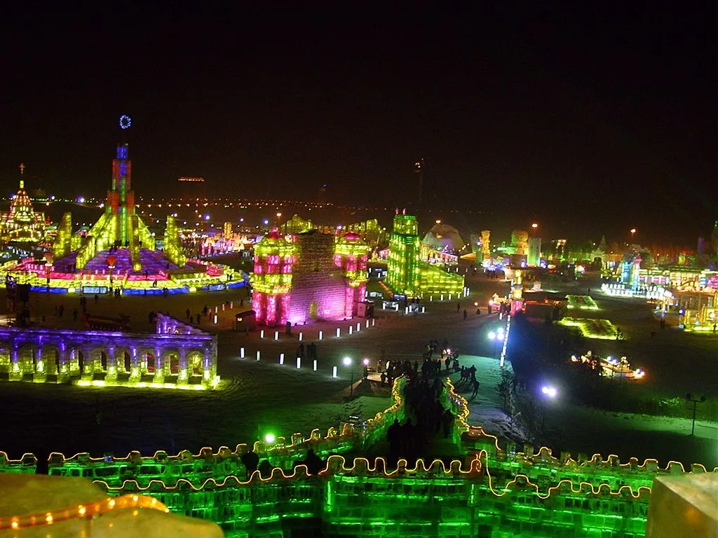 The Harbin Ice and Snow World at night — the 600,000 sqm illuminated ice city lit up in rainbow LED colors, with ice buildings, towers, and a great wall visible in the wide aerial view; the world's largest ice festival, open only in January-March