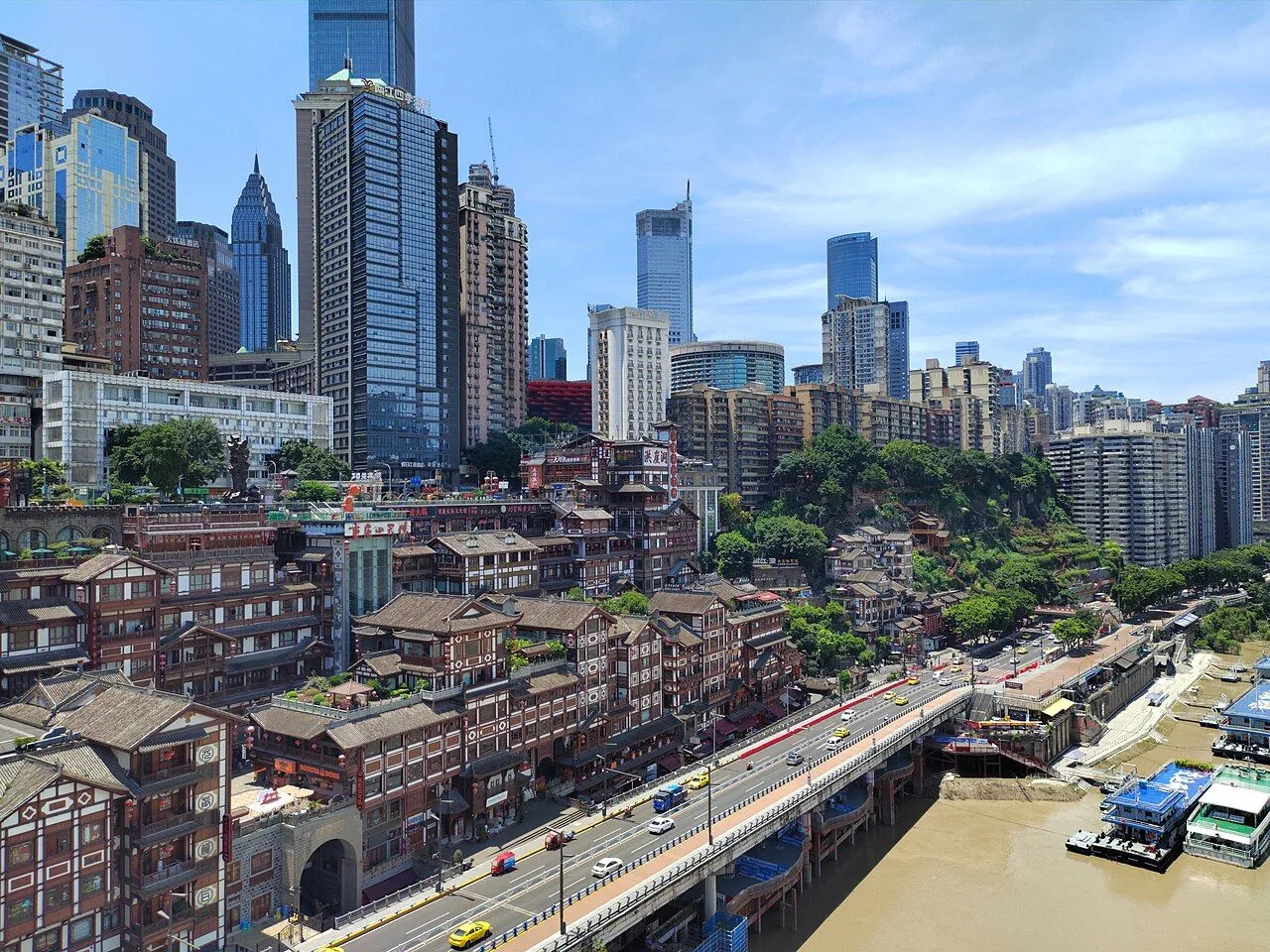 Aerial view of Hongya Cave (洪崖洞) in Chongqing — the 11-story stilted-house complex on the cliff face, the bilingual Chinese-English sign visible, with the modern CBD skyline (Raffles City, IFC) rising in the background above the Yangtze tributary