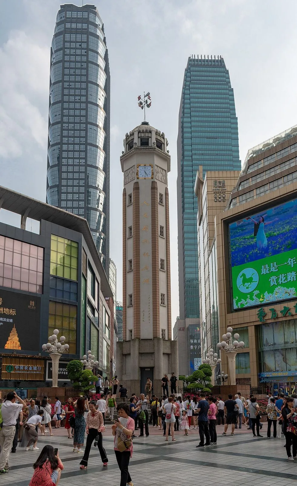 Jiefangbei Square in central Chongqing with the 1947 People's Liberation Monument (解放碑, the 27.5m clock tower in the center) — the heart of the CBD and the original monument that gave the area its name; surrounded by Chongqing's modern skyscrapers and the luxury shopping mall facades