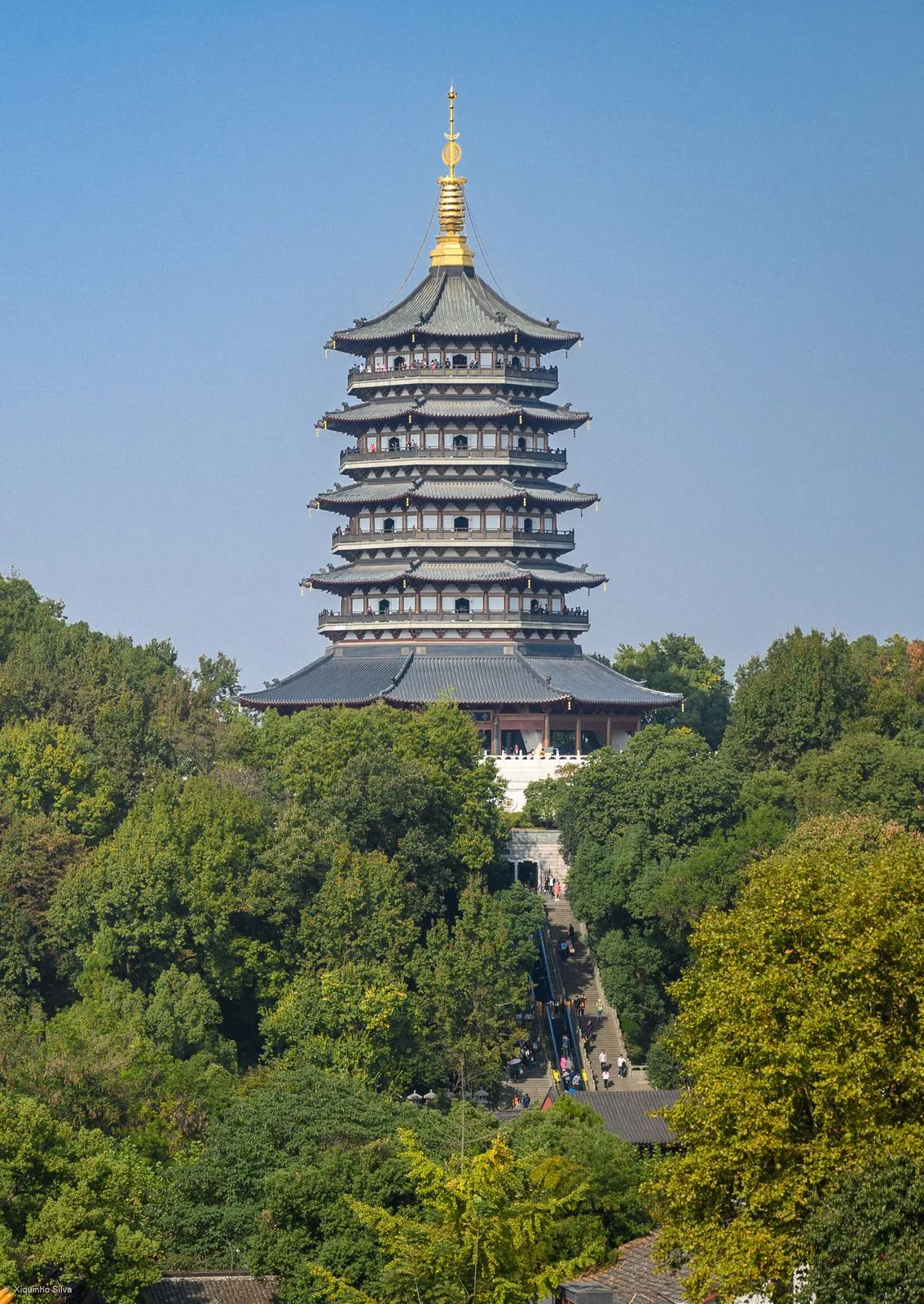 The Leifeng Pagoda at West Lake in Hangzhou, viewed from below against a clear blue sky — a 5-story octagonal pagoda with traditional upturned eaves, the gold finial at the top, and visitors visible on the upper viewing balconies, with the entrance pavilion at the base of the hill and the long stone staircase leading up to it