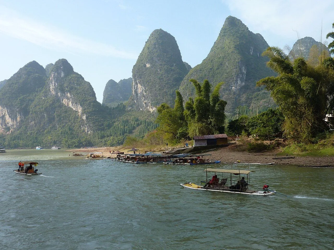 A Li River cruise scene near Xingping — a motorized bamboo raft carrying tourists under a canopy, the iconic karst peaks of Guilin rising behind, and a thatched hut on the riverbank; the most-recognised cruise experience in China, depicted on the 20-yuan banknote