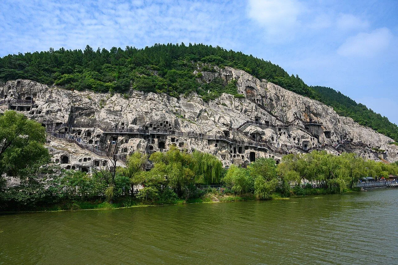 The Longmen Grottoes complex seen from across the Yi River in Luoyang — the white limestone cliff face filled with thousands of cave niches carved over 5 dynasties, willow trees along the riverbank, and the green hills above; a UNESCO World Heritage Site begun in 493 AD during the Northern Wei dynasty