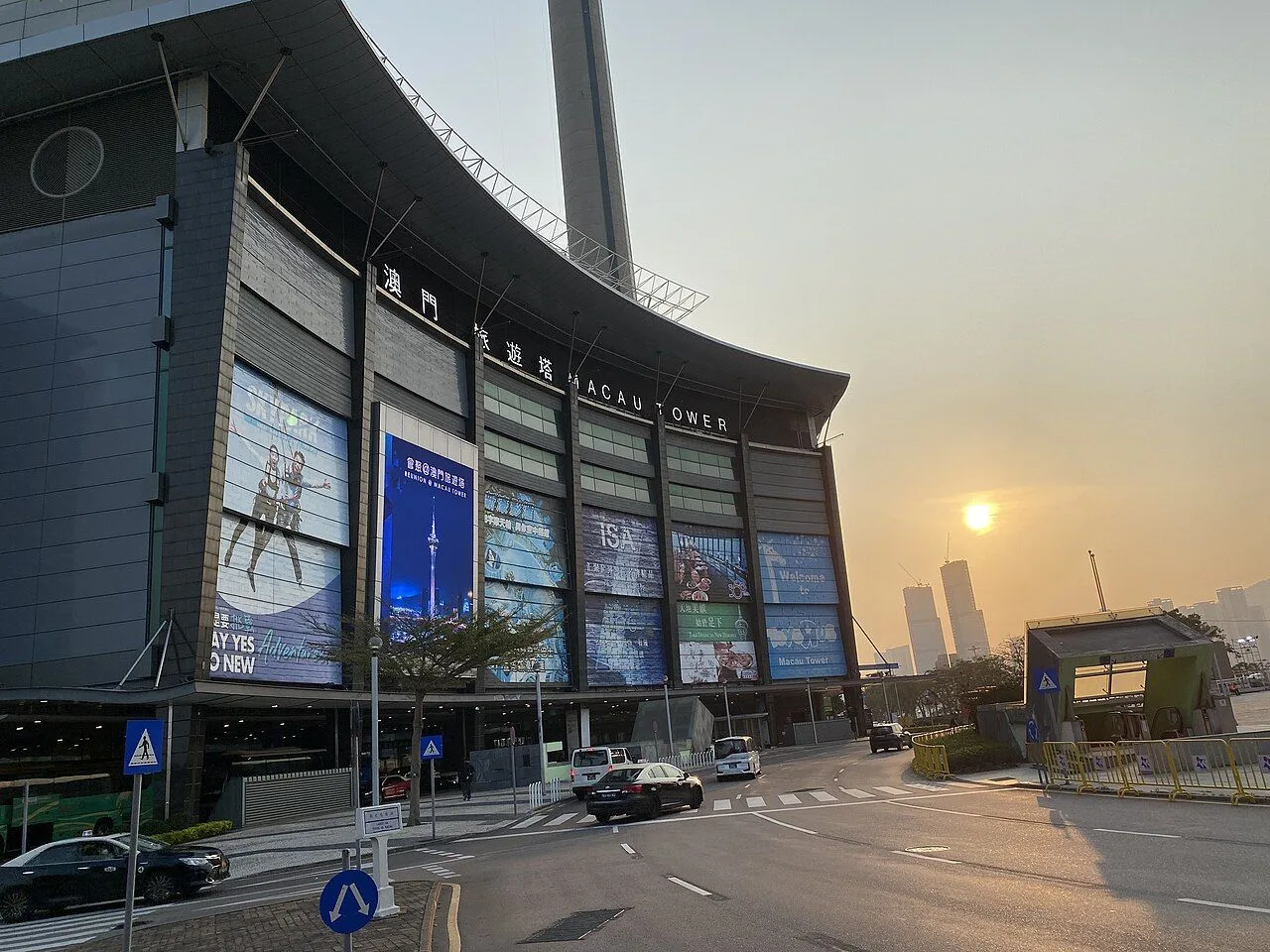 The Macau Tower base at sunset — the 澳門旅遊塔 / MACAU TOWER signage visible on the curved modern facade, with the 338m concrete spire rising behind, the iconic Macau landmark that hosts the highest commercial bungee jump in the world (233m, certified by Guinness)