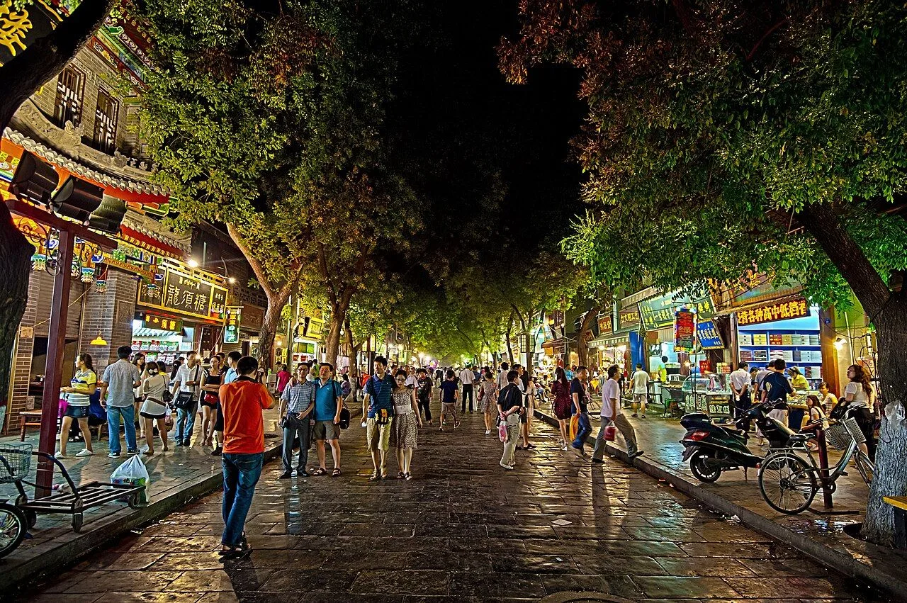 The Xi'an Muslim Quarter at night — a stone-paved street lined with traditional Chinese lanterns, calligraphy shop signs, and Hui Muslim restaurants, crowded with visitors exploring the 500-year-old neighborhood adjacent to the Drum Tower. The bazaar stretches for about 500m along Beiyuanmen Street