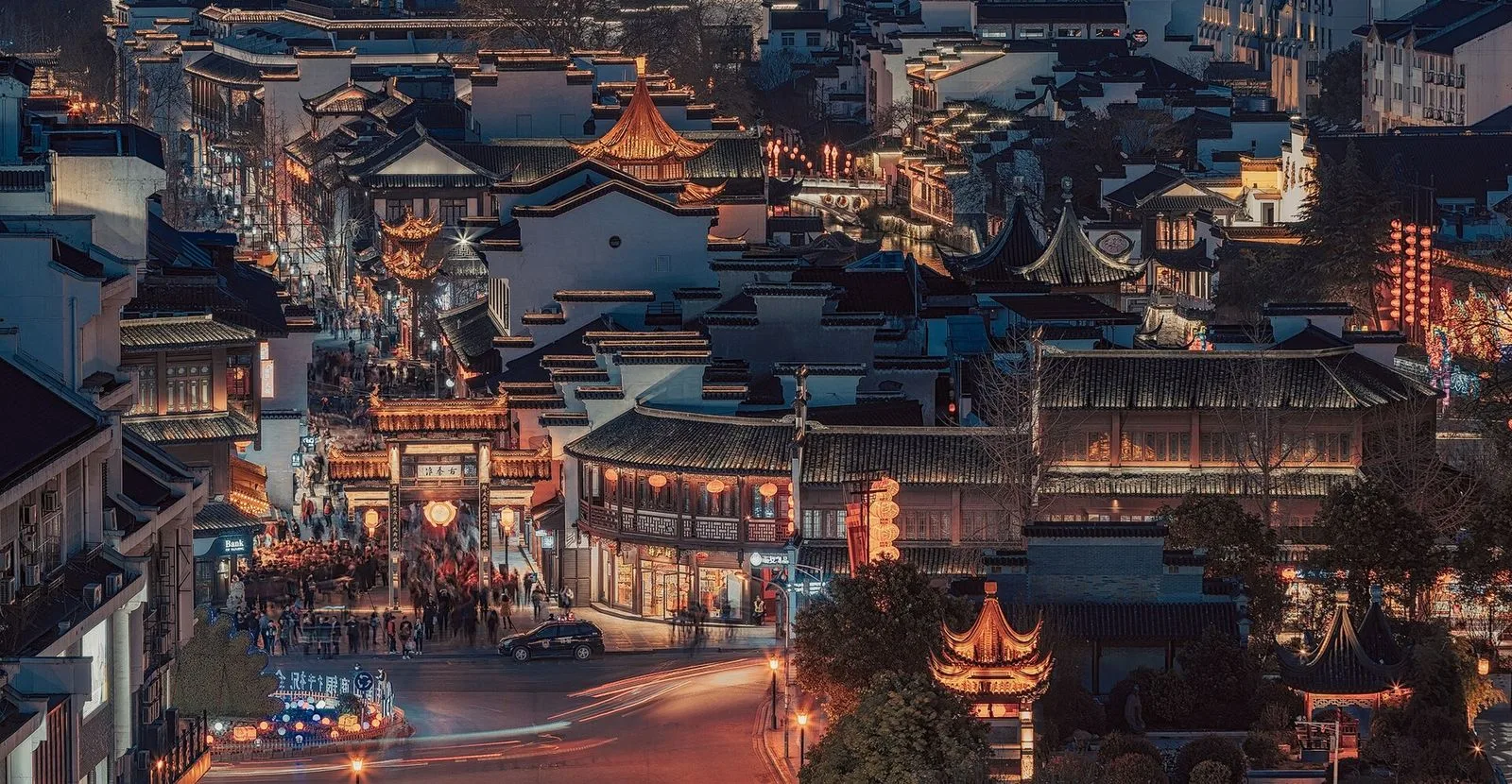 An aerial night view of the Confucius Temple (Fuzimiao, 夫子庙) area in Nanjing — the traditional Chinese rooftops and pagoda lit up with golden lights, the crowds of tourists walking through the illuminated streets, the night view of the Qinhuai River district that draws 30 million visitors a year to the rebuilt Ming-and-Qing-era complex, the heart of southern China’s Confucian heritage and the most atmospheric night-view spot in Nanjing