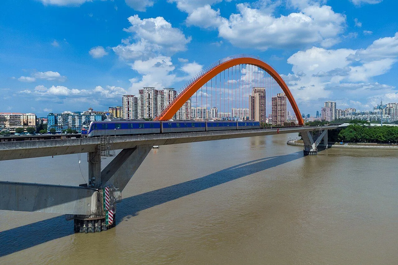 The Jinshazhou Pearl River Bridge, a striking orange tied-arch bridge spanning the muddy waters of the Pearl River in Guangzhou, with a blue and white metro train crossing the deck and the residential skyline of Liwan District in the background under a partly cloudy sky
