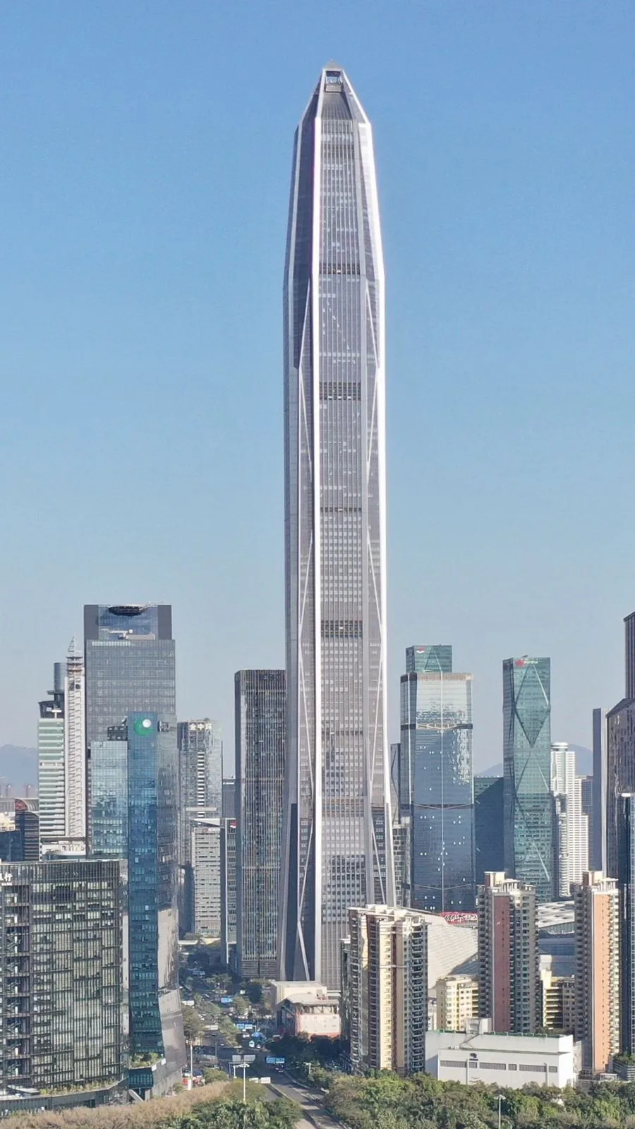 Vertical panoramic view of the 599m Ping An Finance Centre in Shenzhen showing the full height of the tapered glass tower from base to crown against a clear blue sky, with the surrounding CBD skyscrapers and distant green mountains visible at the bottom of the frame