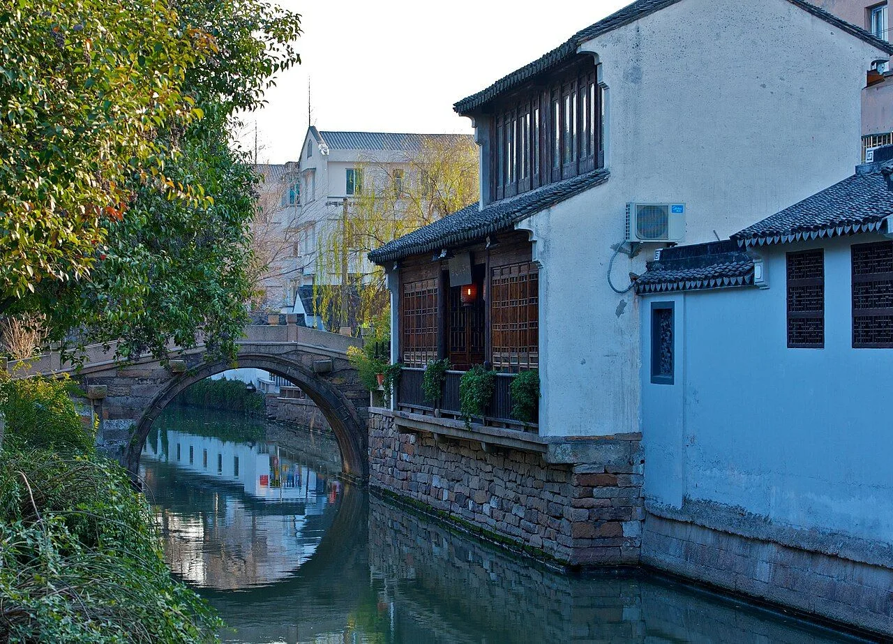 A stone arch bridge over a Pingjiang Road canal — a single-span granite bridge in classic Jiangnan style, with whitewashed traditional houses and weathered brick walls reflected in the still canal water, the quiet riverscape that has defined this Suzhou street for over 1,600 years