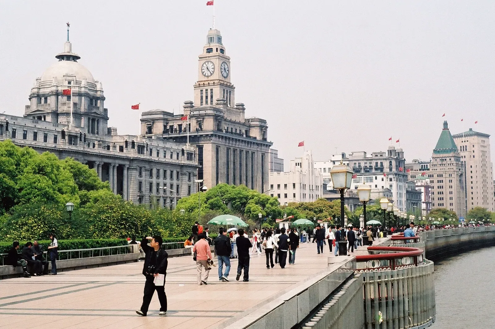 The Bund promenade in Shanghai on a busy day, with the early 20th-century European-style heritage buildings (clock tower, domes, and gold detailing) lining the street and crowds of tourists walking along the riverside walkway facing the Huangpu River