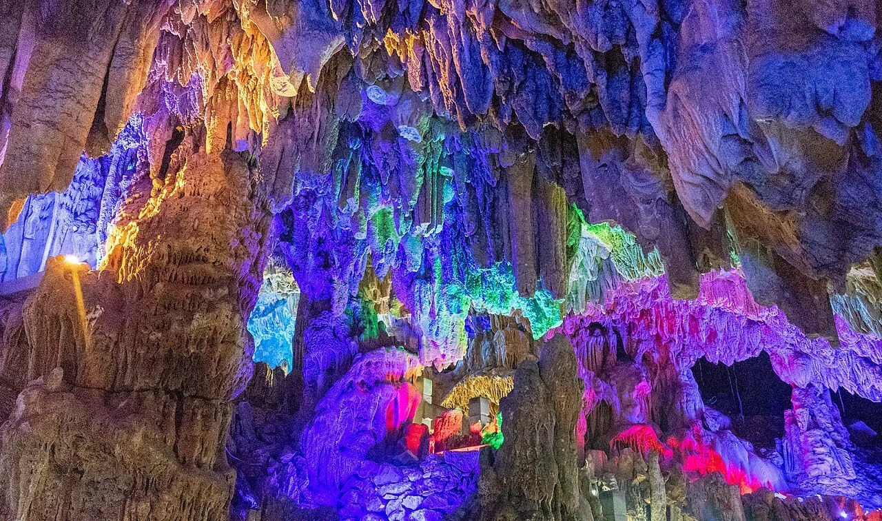 The main hall of Reed Flute Cave (芦笛岩) in Guilin — a 270-million-year-old karst cave illuminated with multicolored LED lights that reveal a forest of stalactites and stalagmites, with formations dripping in electric blue, purple, gold, and magenta the full height of the 18m chamber, the most spectacular room in this 240m-deep cave system