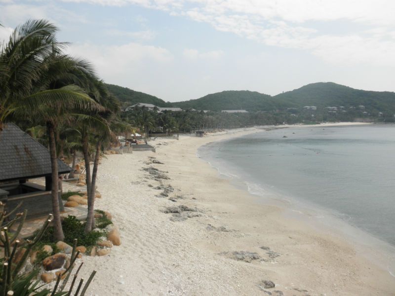 A palm-fringed crescent beach on Yalong Bay (亚龙湾) on Hainan Island — soft white sand, shallow turquoise water, and the green tropical mountains of the Sanya hinterland in the background, the most-celebrated 7.5km beach on the south coast of Hainan and home to a dozen international luxury resorts including the Marriott, Ritz-Carlton, and Pullman