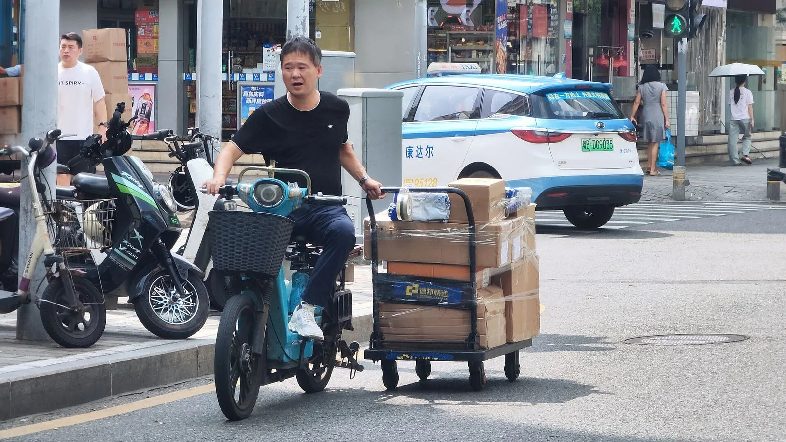 A delivery man on an electric scooter pulling a heavy cart of packages along a Huaqiangbei street, with a blue and white BYD e5 electric taxi in the background and a Huawei store visible — the everyday working scene of the district, with hundreds of delivery riders and shopkeepers moving components, phones, and parcels through the streets every day