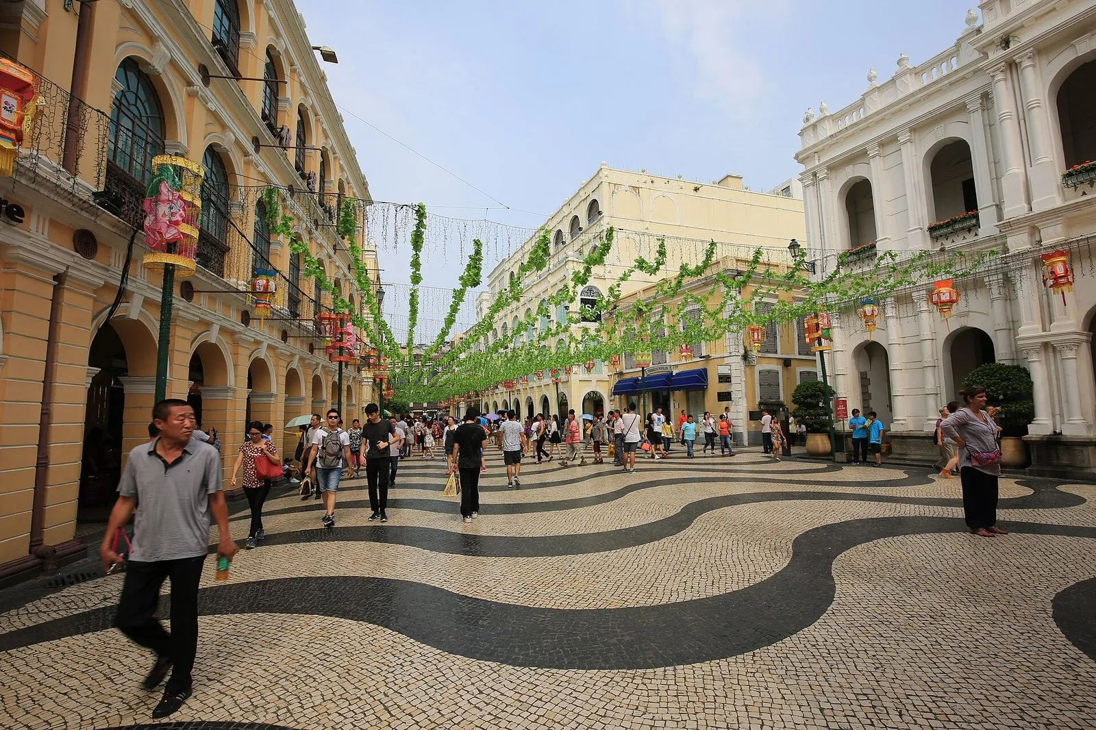 The wave-pattern black-and-white Portuguese pavement of Senado Square (议事亭前地) in Macau — the iconic calçada portuguesa design laid in 1994, surrounded by neoclassical colonial buildings painted in yellow and white with green shutters, the central public square of the UNESCO-listed Historic Centre of Macau that has been the city’s gathering place since the 16th century