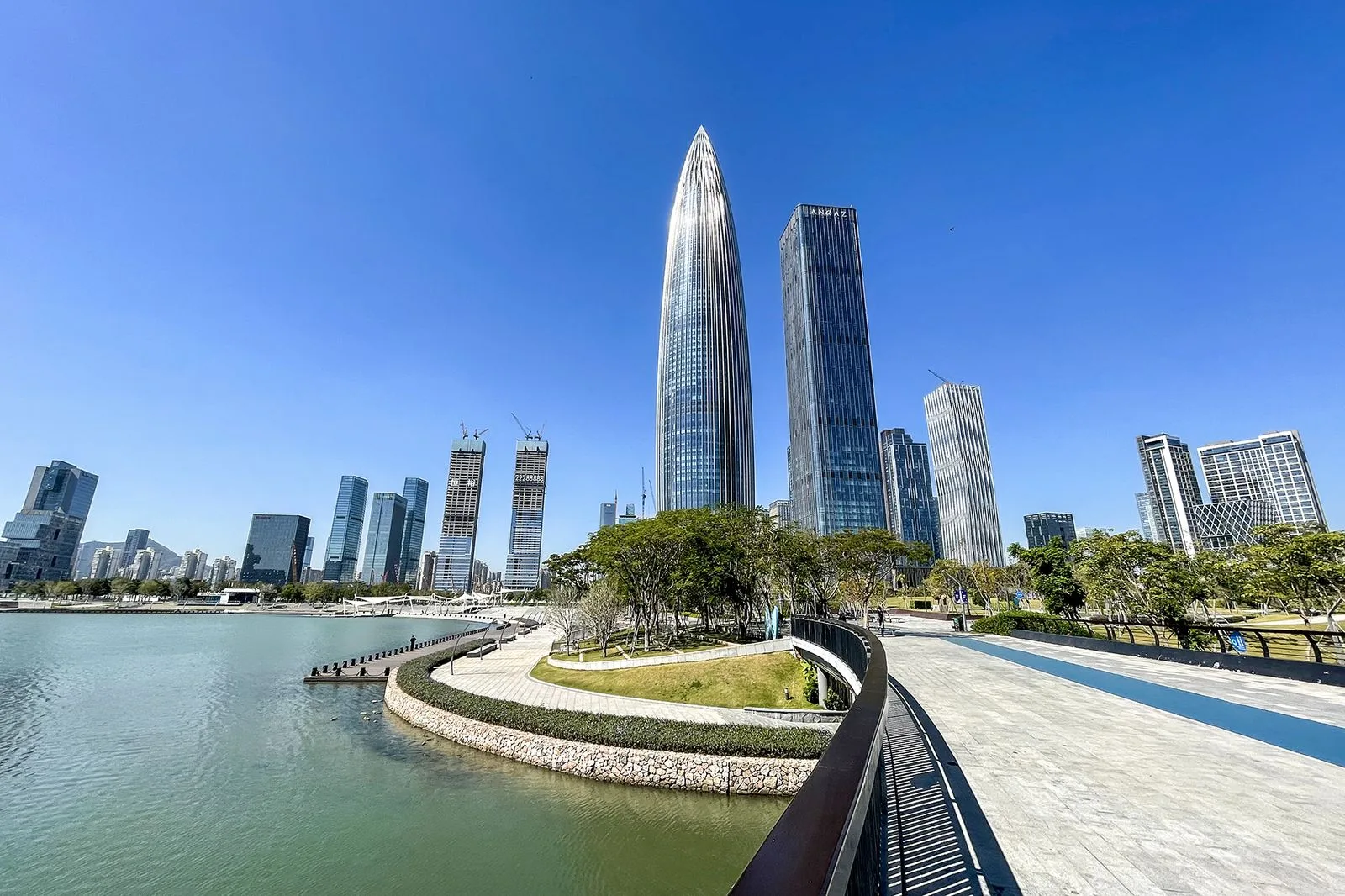 The Shenzhen Bay Park waterfront promenade with the iconic Spring Bamboo shoot tower (春笋大厦, also known as the China Resources Headquarters) and the Tencent Binhai Mansion rising behind, the curved walking path along the bay lined with tropical trees, the best place in Shenzhen to see the contrast between the modern tech skyline of Nanshan District and the natural Shenzhen Bay wetland that hosts migratory birds each winter