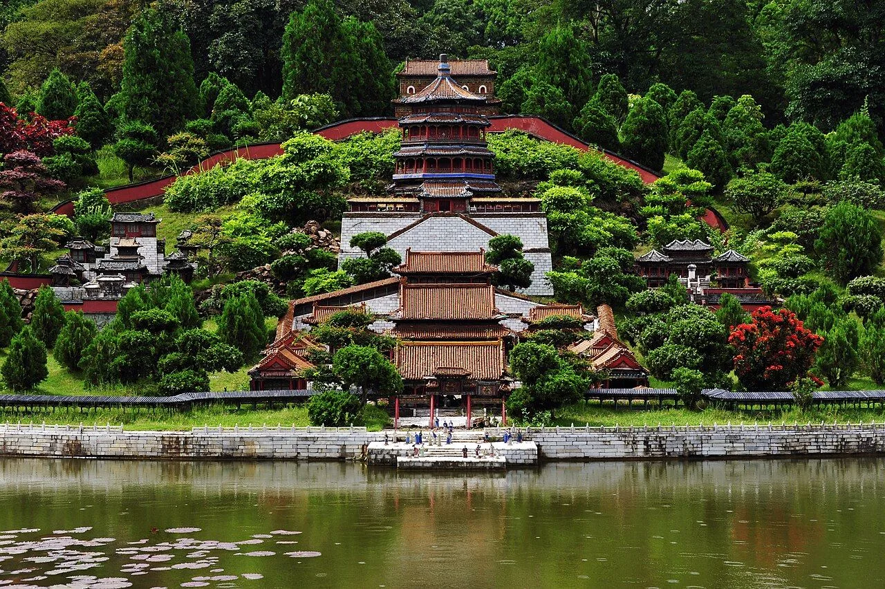 A 1:15 scale miniature replica of the Forbidden City in Splendid China Folk Culture Village Shenzhen, the multi-tiered imperial palace with golden roofs and red walls set against a hillside of green trees, reflected in the lake in the foreground