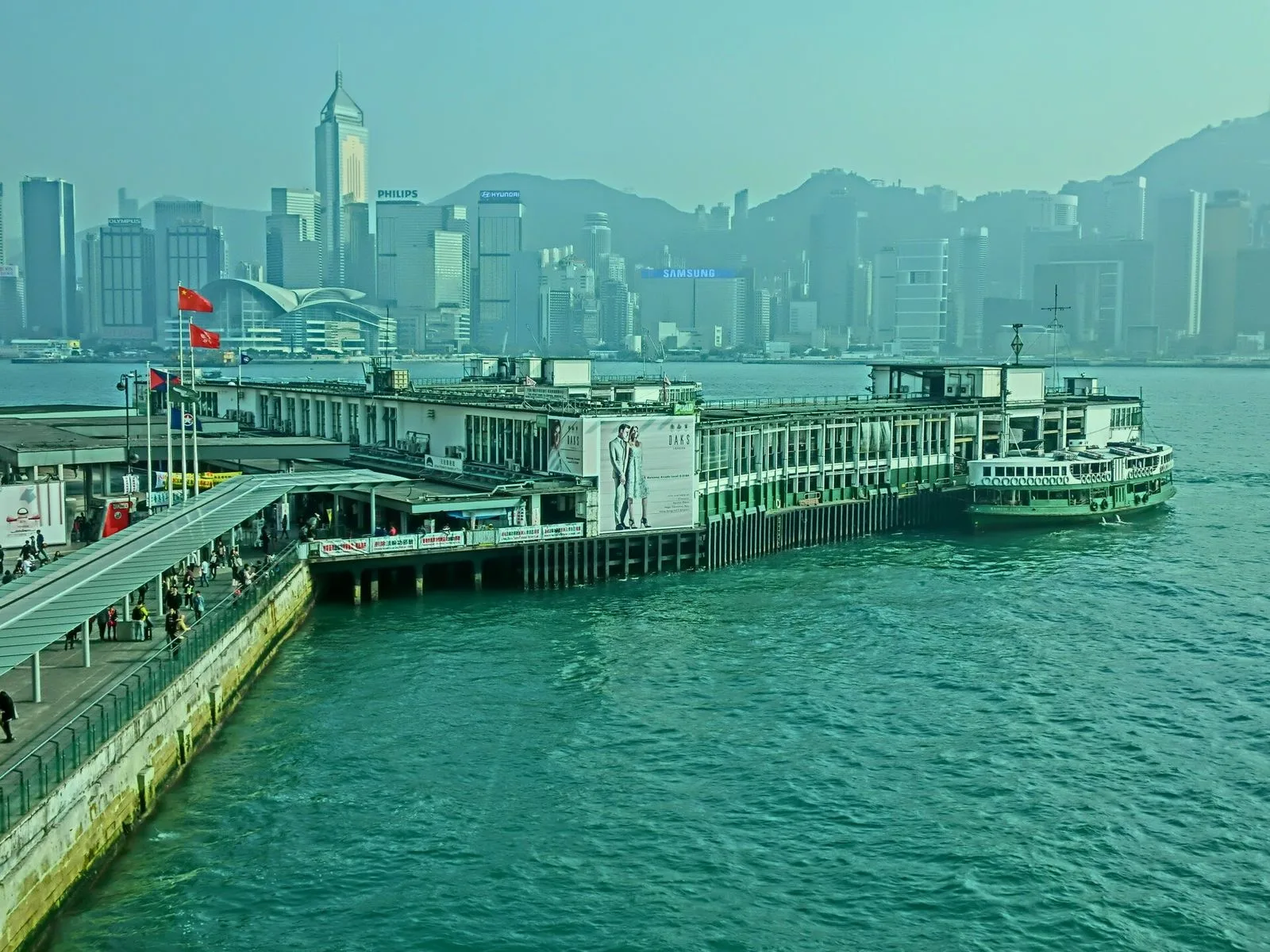The Tsim Sha Tsui Star Ferry Pier in Hong Kong — the green-and-white double-deck ferry pier with the iconic Pier-7 sign, the Hong Kong Convention and Exhibition Centre and the H&M and SAMSUNG-advertised Wan Chai waterfront visible across Victoria Harbour, the 130-year-old ferry service that still carries 60,000 passengers a day across the harbor for just HK$4 per crossing