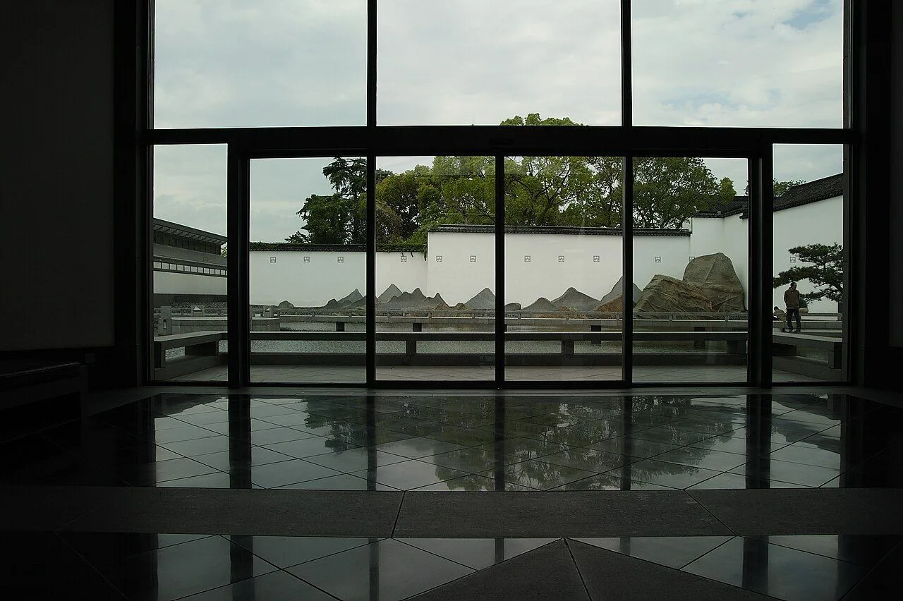 The iconic view from inside the Suzhou Museum: a floor-to-ceiling window perfectly frames the central courtyard with its white walls, dark tile roofs, I.M. Pei's rock garden, and reflecting pool, demonstrating the "borrowed scenery" concept in modernist form
