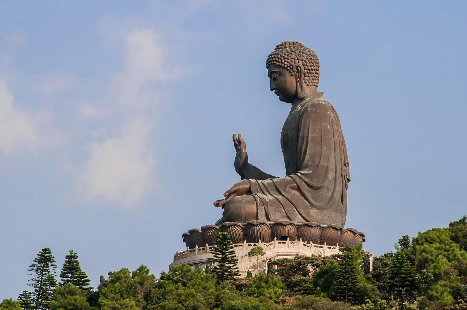 The Tian Tan Buddha (天壇大佛) in profile — the half-face view of the 34m-tall bronze Buddha statue showing the Buddha’s serene features, the right hand raised in Abhaya mudra (the gesture of fearlessness and benediction), the curling hair details and elongated earlobes, the bronze statue cast in 1993 by the Jiangsu Wuxi Jincheng Arts and Crafts Factory and sitting on a three-tiered altar atop Mount Muk Yue on Lantau Island
