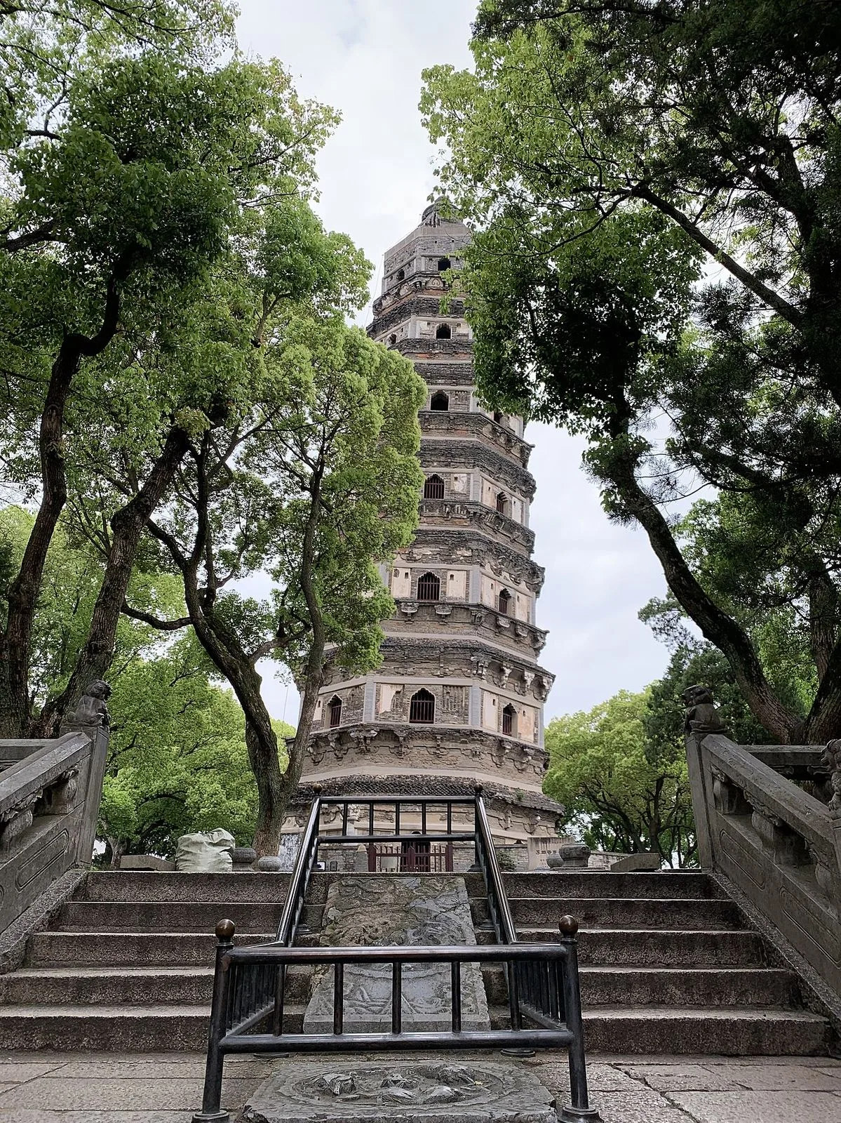 The 47-meter leaning Yunyan Pagoda at the top of Tiger Hill, Suzhou, a 7-story brick pagoda tipped 2.34 degrees from vertical — built 961 AD and leaning for over 1,000 years, framed by tall trees and viewed from the base of the stone steps leading up to it