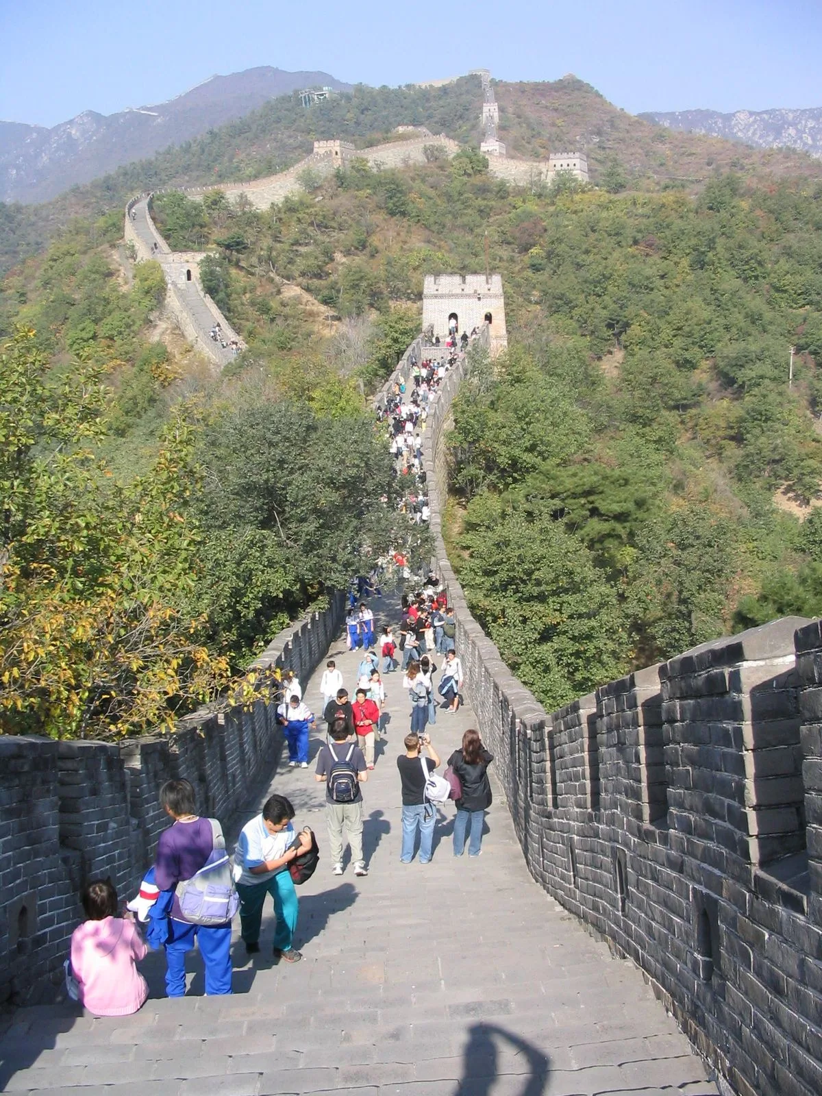 Tourists walking on the restored walkway of Mutianyu Great Wall, with the wall snaking along forested mountain ridges into the distance, watchtowers visible at intervals, and a clear blue sky overhead — the everyday visitor experience with enough space to walk at your own pace, unlike the shoulder-to-shoulder crowds at Badaling