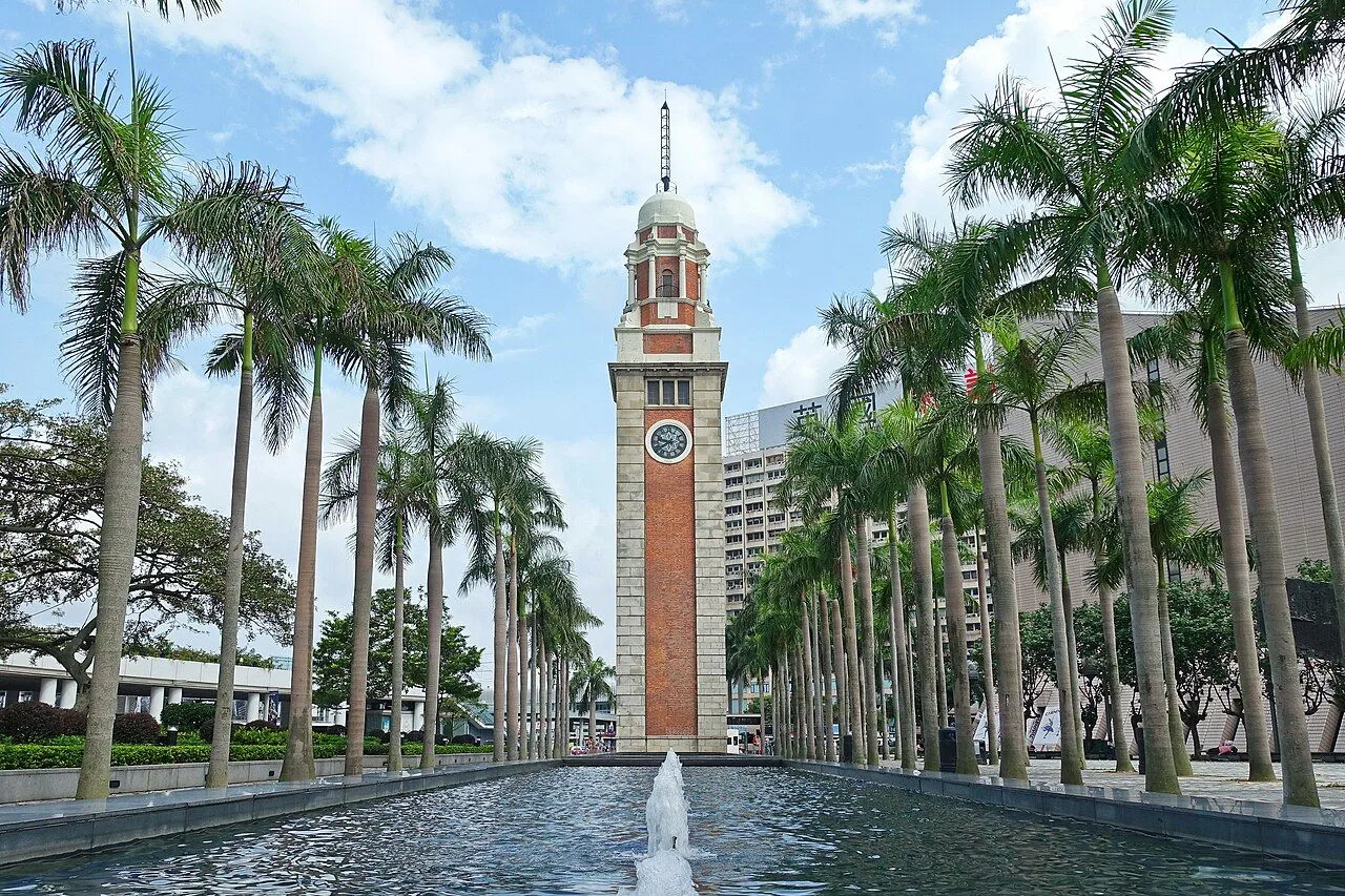 The 44-meter Former Kowloon-Canton Railway Clock Tower at Tsim Sha Tsui, built 1915, the only surviving piece of the old Kowloon Station, framed by two rows of tall palm trees and reflected in the long pool in front, against a partly cloudy sky
