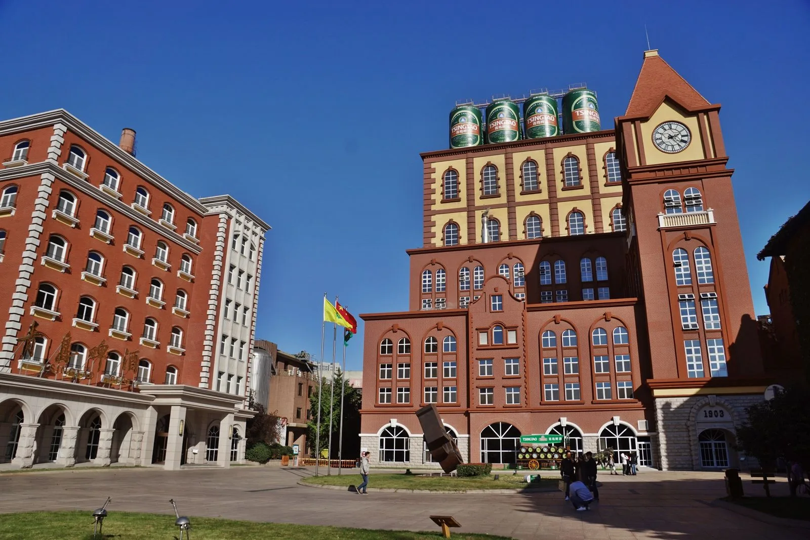 The red-brick Tsingtao Brewery complex in Qingdao — the German colonial-era brewery buildings with the distinctive 1903 Germanic red-brick facade, the green Tsingtao beer cans stacked outside for visitors to photograph, the original brewery that was founded by the Anglo-German Brewery Co. in 1903 in Qingdao (then a German colony called Tsingtau) and still produces beer on the same site, the most-visited brewery museum in China drawing 800,000 visitors a year