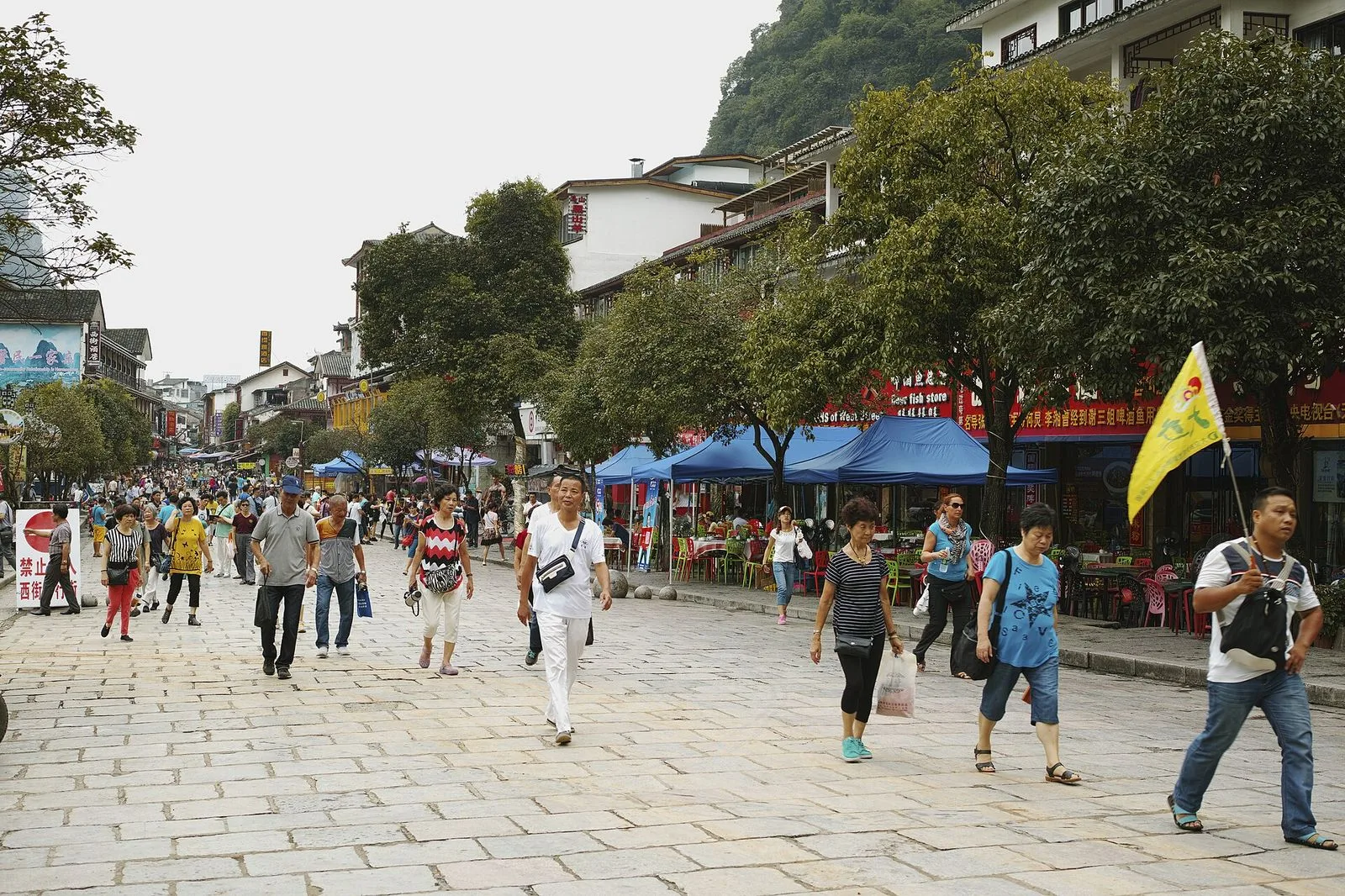 The iconic entrance of West Street (西街) in Yangshuo — the traditional Chinese gate on the right with the English name "West Street Xijie Foreign Department Store" written in cursive gold letters, the dramatic karst peaks of Guangxi rising in the misty background, the stone-paved pedestrian street stretching into the distance, the 1,400-year-old street that has been the foreigner hangout in southern China since the 1970s and is the most-visited street in Guangxi