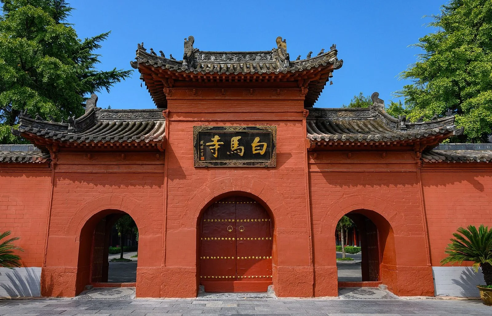 The iconic red gate of the White Horse Temple (白马寺, Baimasi) in Luoyang — the golden Chinese characters 白马寺 (White Horse Temple) on the black plaque above the central red door, the traditional Chinese roof with upturned eaves and the three-arched entrance, the temple gate that has stood since the temple was founded in 68 AD as the first Buddhist temple in China, brought by the Indian monks Kasyapa Matanga and Dharmavarma