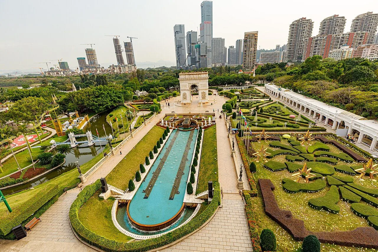 A 1:3 scale replica of the Arc de Triomphe at the entrance of Window of the World, Shenzhen, with the perfectly manicured French garden, geometric topiary, a long ornamental reflecting pool with fountains, and the Shenzhen skyline of modern skyscrapers rising behind the park