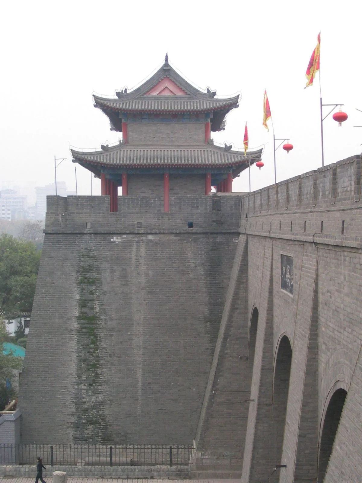 A corner watchtower of the Xi’an city wall — the imposing multi-tiered tower with the traditional Chinese roof and the battlement extending into the distance, the red and yellow flags flying above the brick walls, the wall built in 1370 during the Ming dynasty under the command of Zhu Yuanzhang and still standing as the best-preserved Chinese city wall after 600+ years