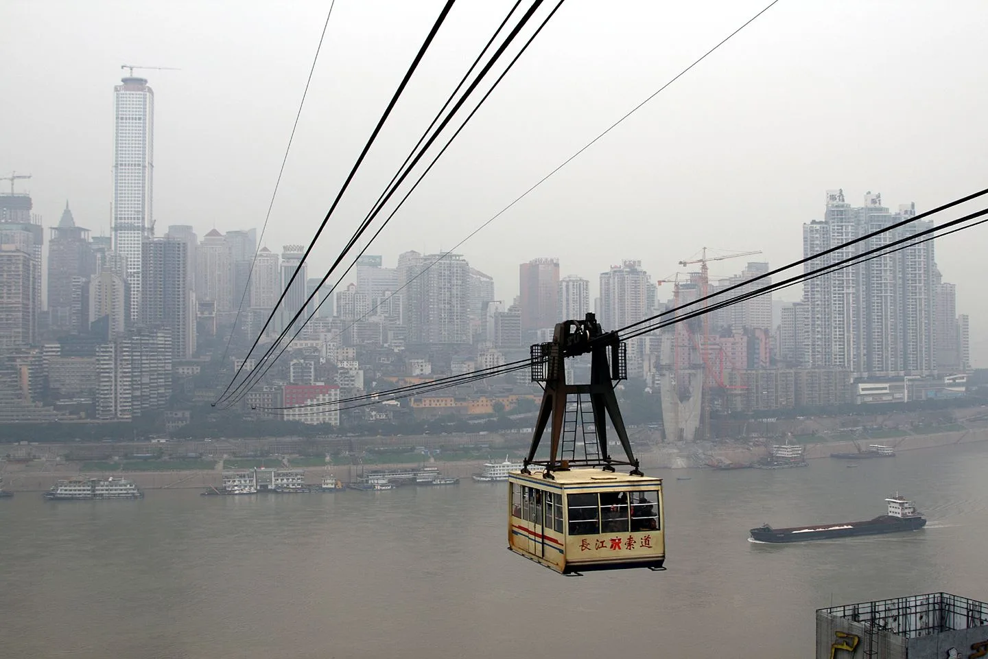 The iconic yellow cable car cabin of the Chongqing Yangtze River Cableway (长江索道) mid-flight over the Yangtze River — the small yellow cabin with the Chinese characters 长江索道 painted in red on the side, the steel cable attaching to the top of the cabin, the Chongqing skyline and boats on the Yangtze in the background, the cable car that crosses the 1,166m distance between Yuzhong and Nan’an districts at 40m above the water in just 5 minutes