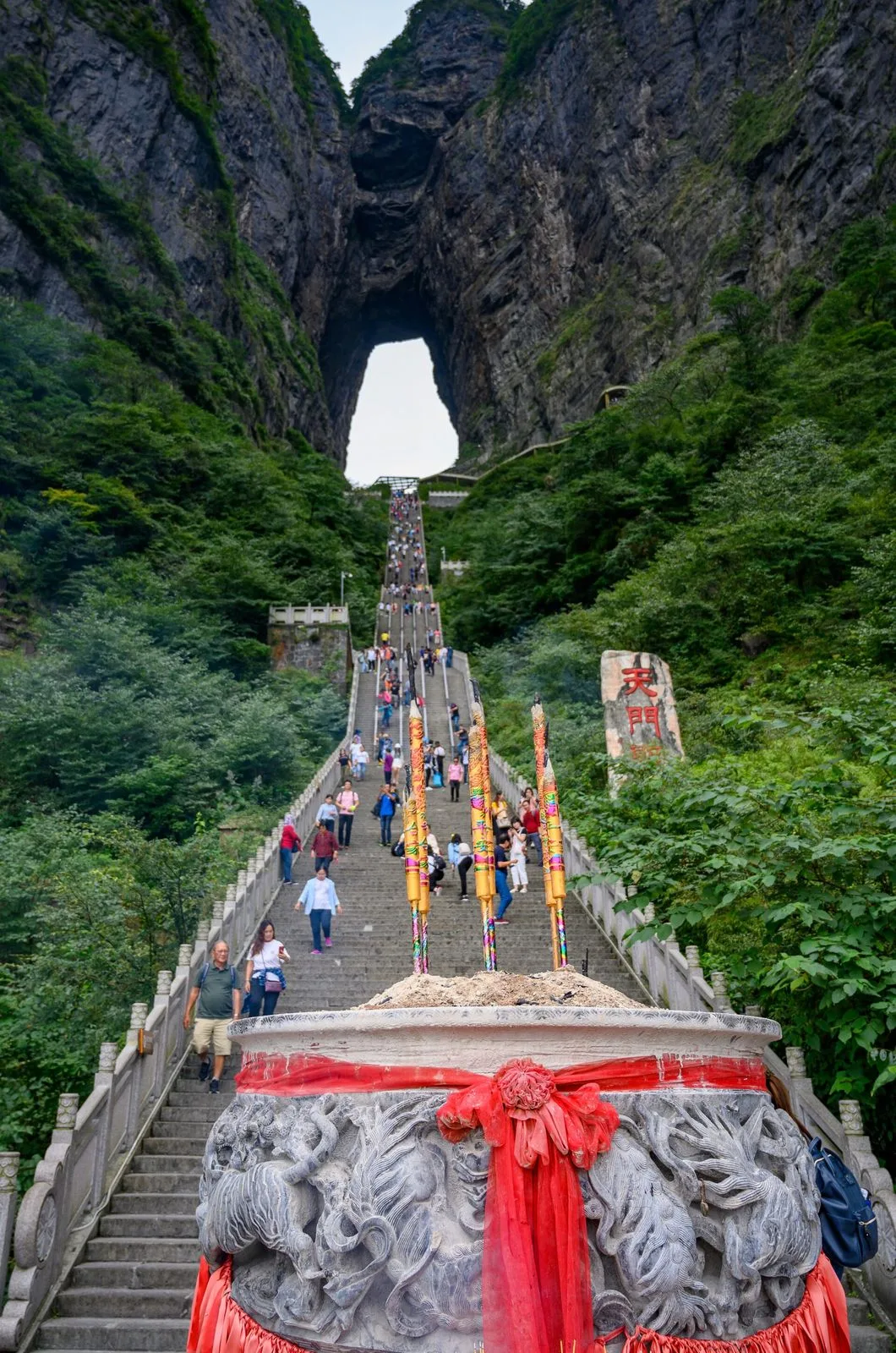 The iconic view up the 999-step Heavenly Stairway to the Tianmen Cave (Heaven’s Gate) on Tianmen Mountain in Zhangjiajie — the stone steps climbing steeply through the narrow valley, the natural arch of the cave at the top, the incense burner in the foreground with the red ribbons, the tourists climbing the steps, the dramatic shot that has become the most-photographed image of Zhangjiajie and the symbol of the mountain that inspired the floating Hallelujah Mountains in James Cameron’s 2009 film Avatar
