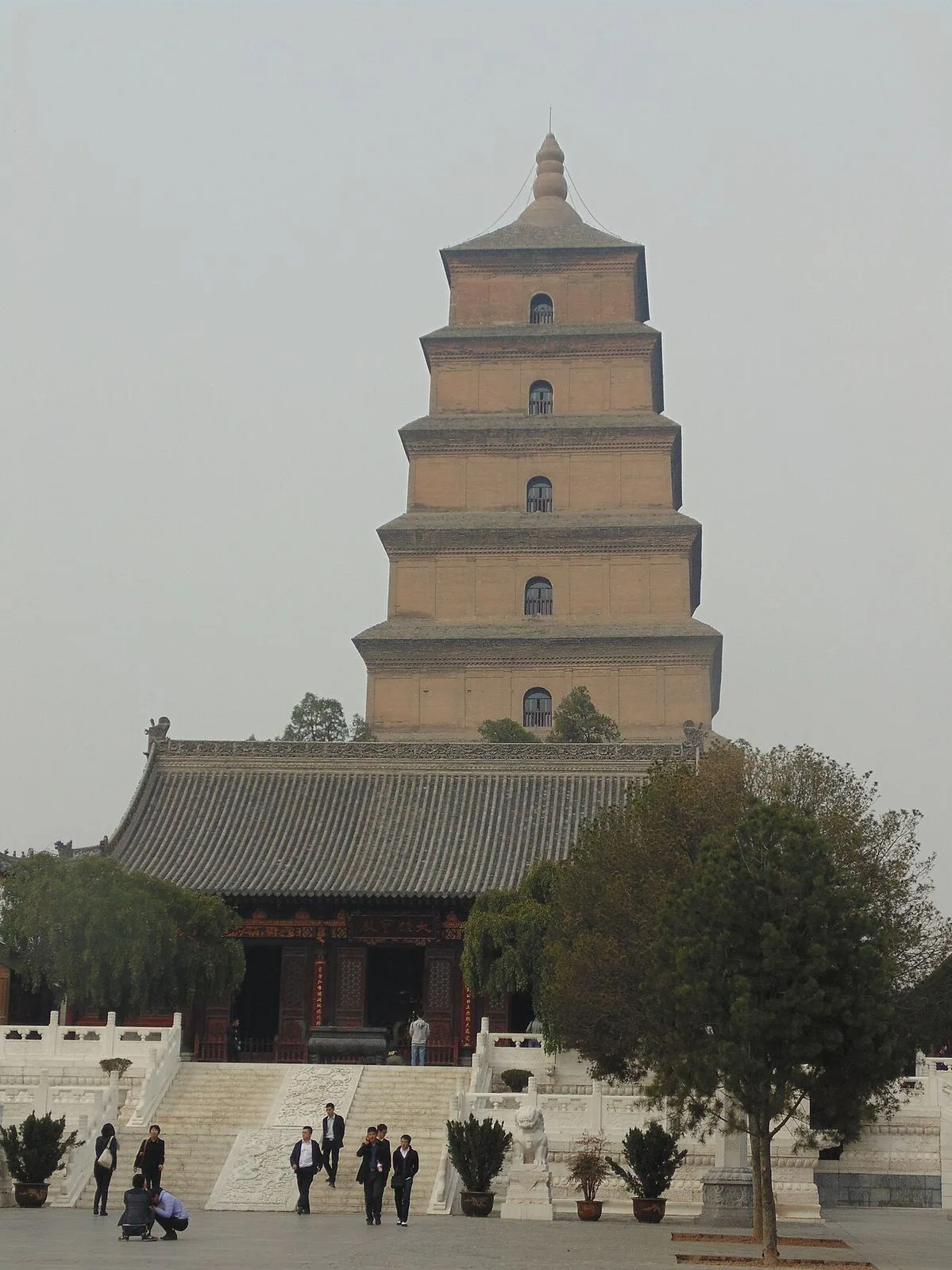 The Big Wild Goose Pagoda towering above the Da Ci'en Temple main hall in Xi'an, with the traditional grey-tiled temple roof in the foreground and tourists on the stone steps — the iconic skyline silhouette of the city