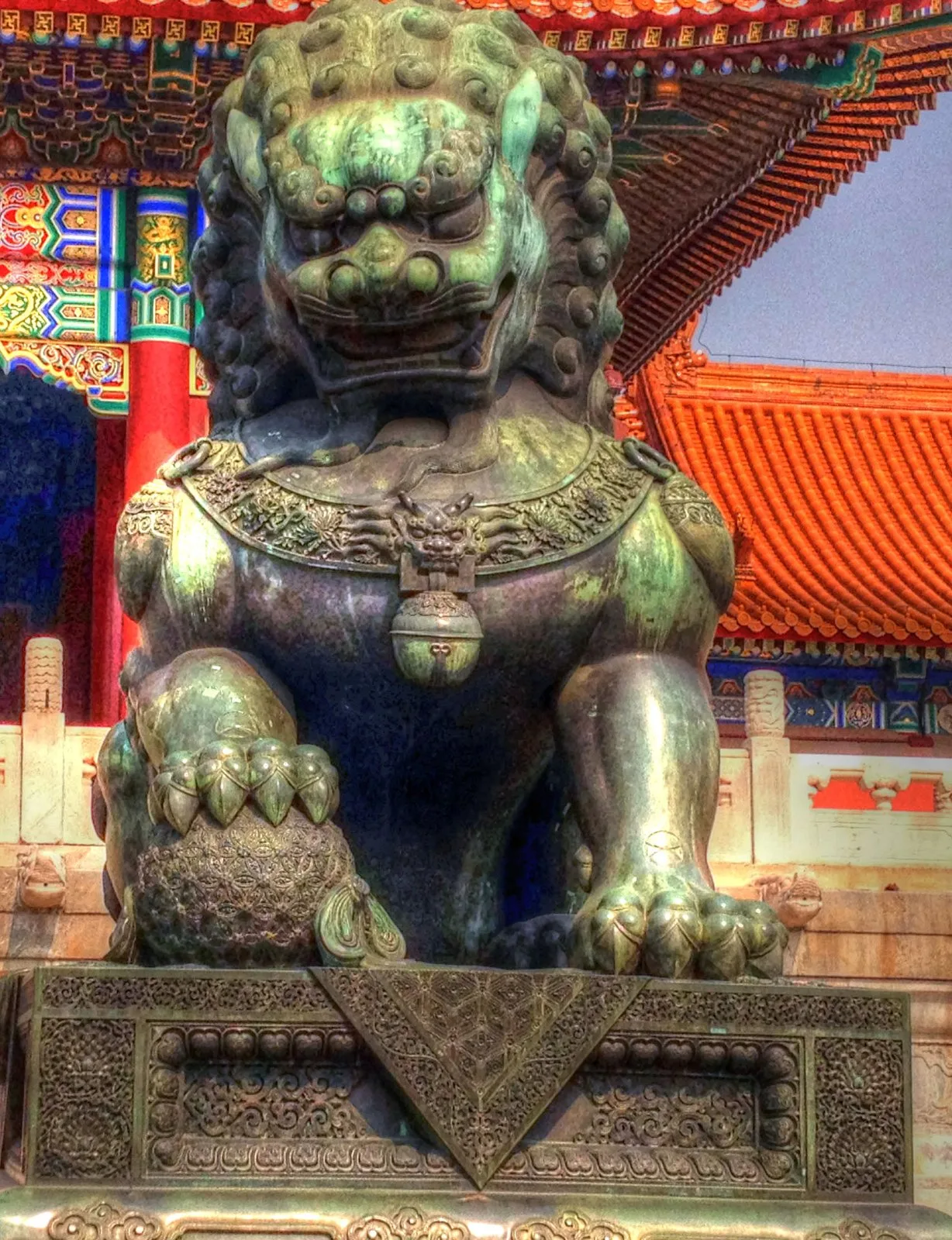 One of the giant bronze guardian lions at the Forbidden City — a Ming-dynasty gilt-bronze sculpture with a green patina and a curled mane, mounted on an ornately carved stone pedestal with paw resting on a decorative embroidered ball, in front of the golden roof tiles of a Forbidden City hall.