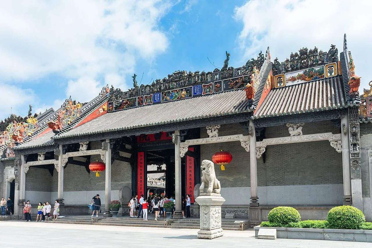 The Chen Clan Ancestral Hall in Guangzhou from a wider angle, with the foreground stone lion, the ceramic-figurine covered roof ridges, the red signboard, and the 11,000+ pieces of ceramic, stone, and wood carving visible across the facade