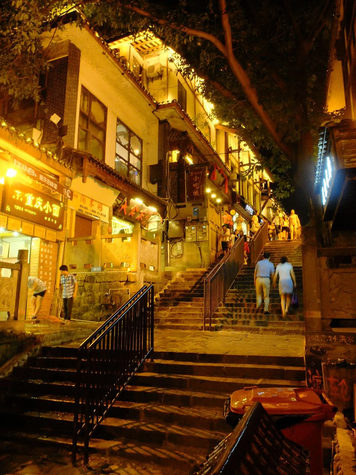 Night view of Ciqikou Ancient Town showing the illuminated stone staircase climbing the hillside, with traditional shopfronts lit by warm yellow lights and visitors walking up and down the steps