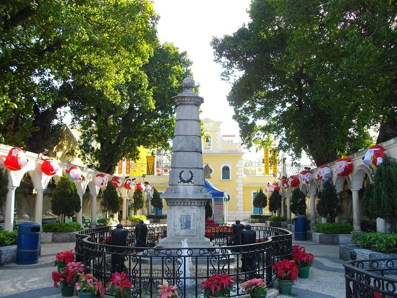 The Coloane Village square in Macau, with the central stone obelisk (the Bom Menino memorial), Santa Claus Christmas decorations strung between the arcade columns, potted poinsettias, and the yellow St. Francis Xavier Chapel in the background