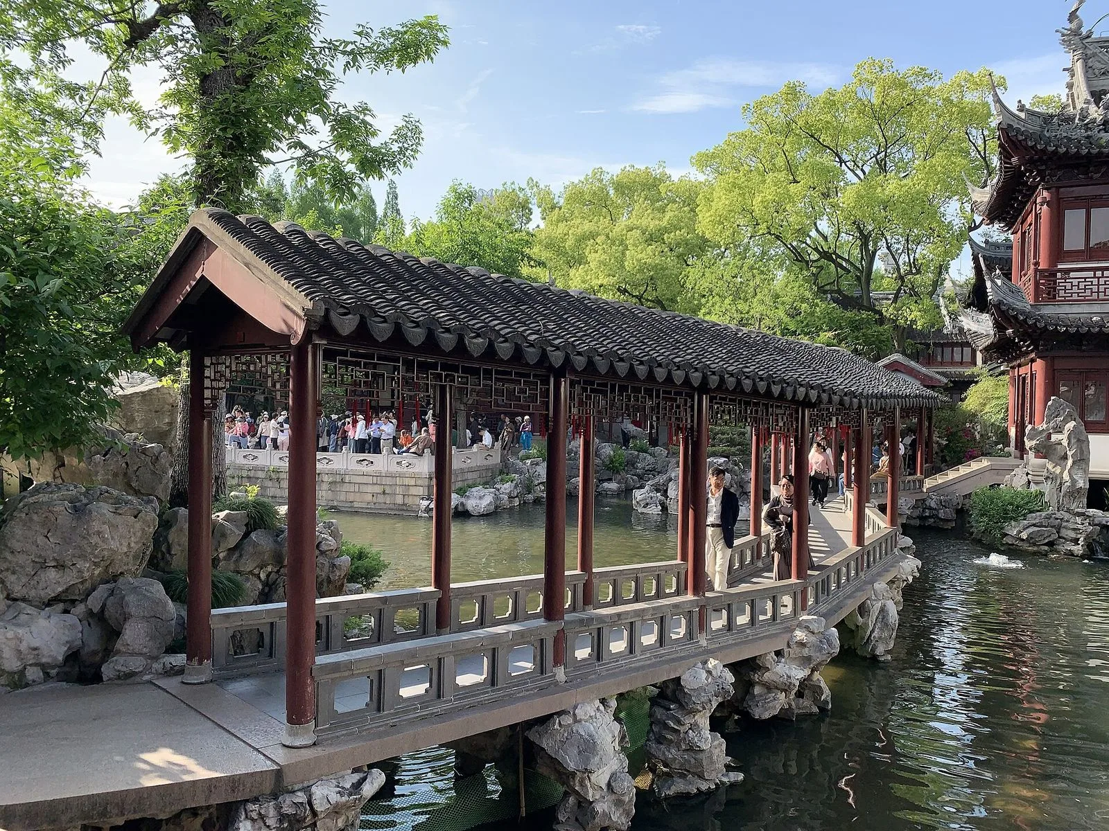 A view of the covered bridge in Yu Garden on a sunny day, with two visitors — a man in a dark blazer and a woman with a scarf — standing in the center of the bridge and posing for photos, the pond visible below and the traditional red-columned pavilions of the garden rising in the background. The bridge is the most-photographed spot in the garden and a Shanghai landmark